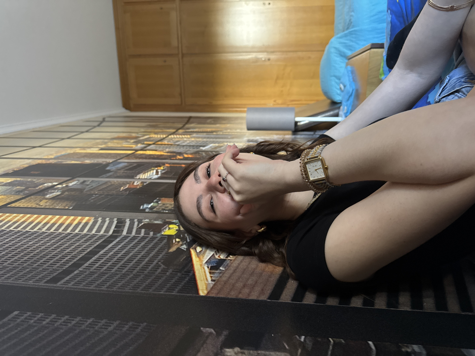 A young woman lies on a floor with a photorealistic city grid design, covering her mouth with her hand. She wears a black top, a gold watch, and a gold bracelet. A large wooden cabinet is visible in the background along with a blue blanket and exercise equipment. The location in Mirandela, Portugal is not identifiable from the image.