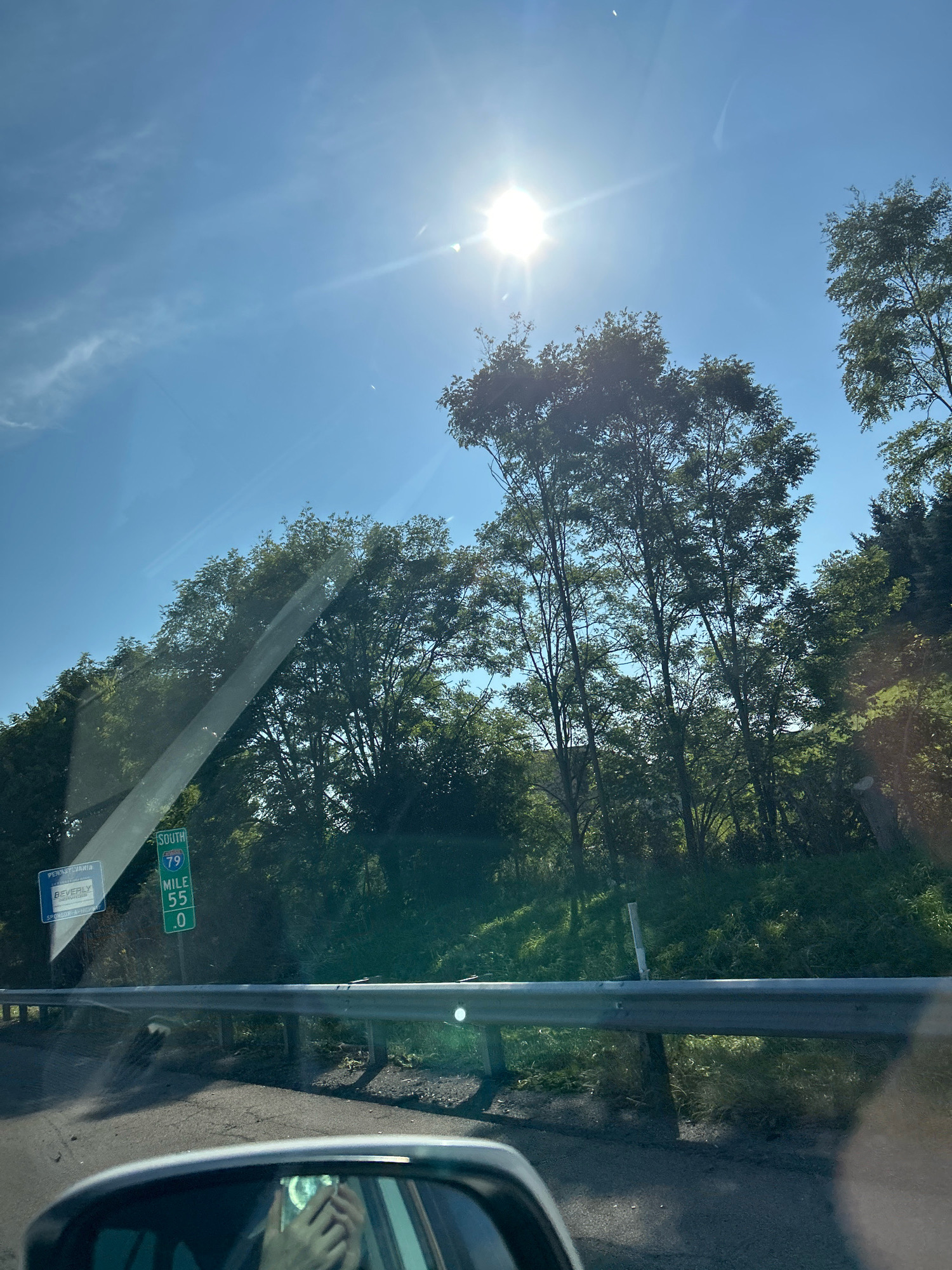 The image captures a sunny day in Collier Township, Pennsylvania, United States of America. The viewpoint is from inside a vehicle, looking forward and slightly upwards. The sky is a clear, bright blue with the sun prominently featured, casting a lens flare across the upper portion of the frame. Several trees with dense green foliage are visible, some reaching towards the sky, creating a dappled light effect on the road below.
On the side of the road, a reflective guardrail runs horizontally. Mounted on posts along this guardrail are road signs. One prominent sign indicates "SOUTH 79", with "MILE 55" and ".0" displayed below. To the left of this sign is a smaller blue sign that reads "PENNSYLVANIA" at the top, followed by "BEVERLY" and "SPONSORED BY".
In the lower right of the image, a portion of the vehicle's interior is visible, including the side mirror, which reflects the driver holding a smartphone. The road surface appears to be asphalt, with some wear and tear visible. The overall environment suggests a scenic drive through a wooded area on a clear day.