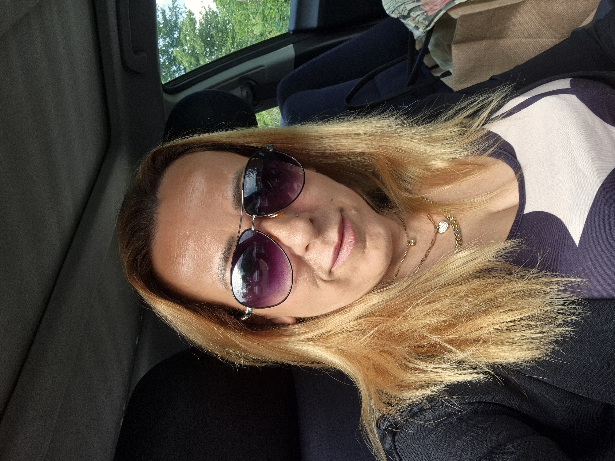 This media file depicts a woman inside a vehicle, likely a car, during the daytime. The woman has long, blonde hair and is wearing dark, aviator-style sunglasses. She has a gold-colored chain necklace with a heart-shaped pendant around her neck. Her lips show a hint of color, and she has a subtle, pleasant expression, almost a slight smile. She is wearing a top with a prominent white design on a dark blue or purple background.
The setting appears to be the interior of a car, with parts of the ceiling, door frame, and dark seats visible. Through the car window in the upper background, bright green foliage from trees can be seen, indicating an outdoor or natural environment. The scene is well-lit, suggesting bright daylight. In the background, behind the woman, a light brown paper bag and a patterned fabric bag are visible. There is no visible text in the image.