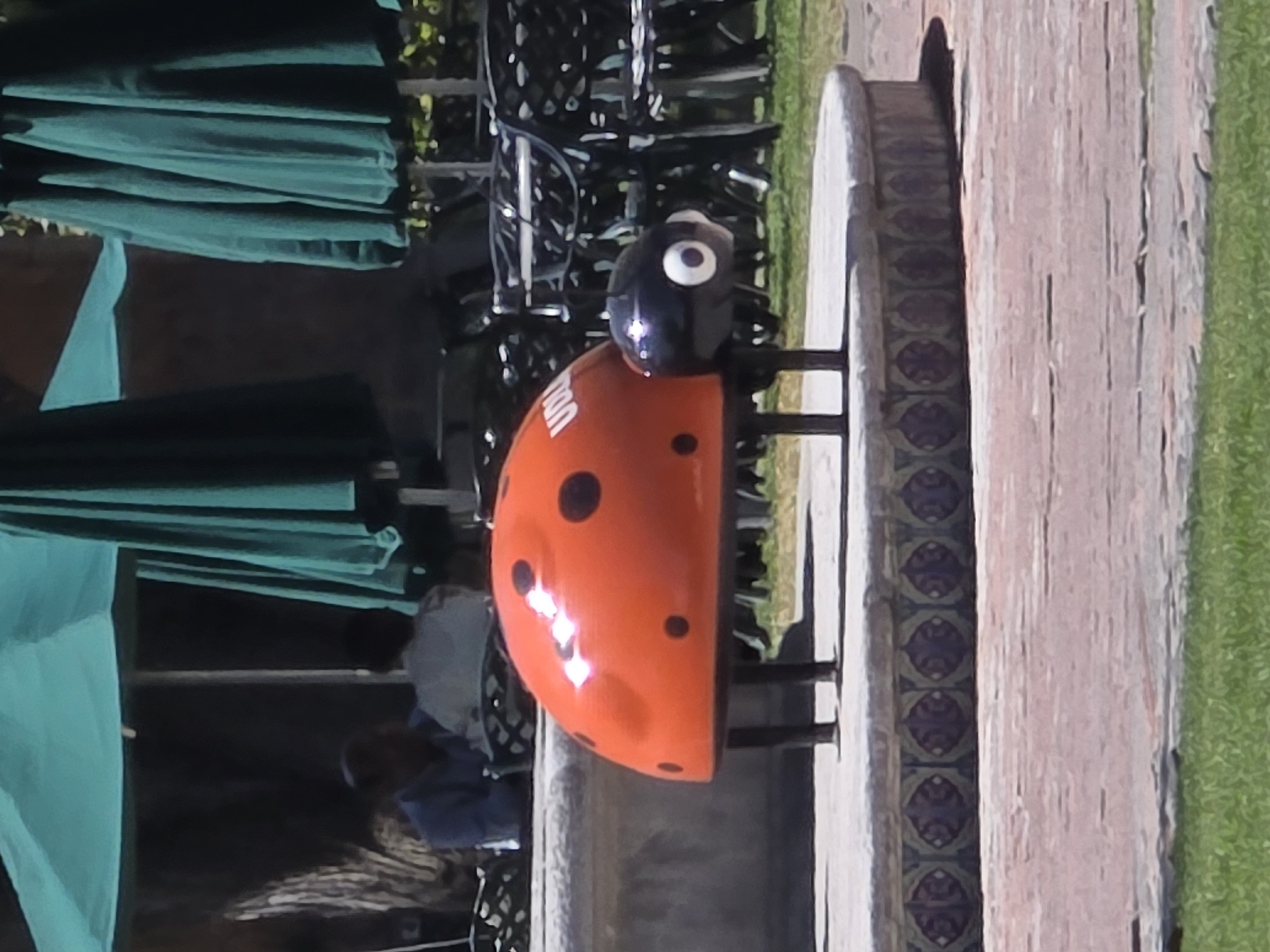 This outdoor scene, located in San Andrés Cholula, Mexico, displays a large, stylized ladybug sculpture prominently positioned on a circular, elevated platform. The ladybug has a glossy orange shell marked with black spots, a black head featuring two large white eyes, and six black legs. Partial white lettering, "UDL," is visible on its orange back. The circular platform is decorated with a repeating pattern of blue, red, and beige ornate tiles. The ground immediately in front of the platform consists of reddish-brown pavers, leading to a strip of green grass in the foreground. In the background, a seating area typical of a café or restaurant patio is observed. This area features multiple dark green patio umbrellas, some appearing open and some folded. Numerous dark-colored metal chairs with intricate lattice designs are arranged around tables. Two individuals are partially visible seated at a table on the left side of the background. One person wears a light blue shirt and faces right, while the other, dressed in light-colored attire, sits with their back mostly towards the viewer, facing left. Blurred foliage and indistinct building structures complete the distant background.
