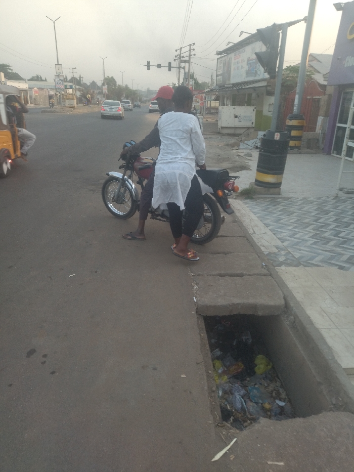 The image shows two people standing next to a motorcycle on the side of a paved road in New Karu, Nigeria. The motorcycle is facing the road, and the two people are standing next to it, with one person appearing to be getting off. The person getting off is wearing a white long-sleeved shirt and appears to be a woman. The other person is wearing a red shirt, and appears to be a man. Both individuals have their backs to the camera. The motorcycle is parked on the sidewalk, which is made of concrete. The sidewalk is next to the road, which is paved with asphalt. There is a large drain on the sidewalk that is filled with trash. The road is wide and straight, and there are buildings on either side of it. The sky is overcast, and the ground is dry. There are several power lines running above the street, as well as one traffic light that is not active. The road is empty of vehicles, except for the motorcycle, a yellow-green vehicle, and a white sedan in the background. The time of day is either early morning or late afternoon, as the sky is somewhat hazy and there are long shadows. There is no vegetation in the scene, and the ground is dry and dusty. The overall scene is a typical urban street in a developing country. There is a sign in the background which advertises a business that sells phones, accessories, and airtime. A trash can on the sidewalk is also visible in the background. The picture appears to be taken from a position on the sidewalk, looking down toward the street.
