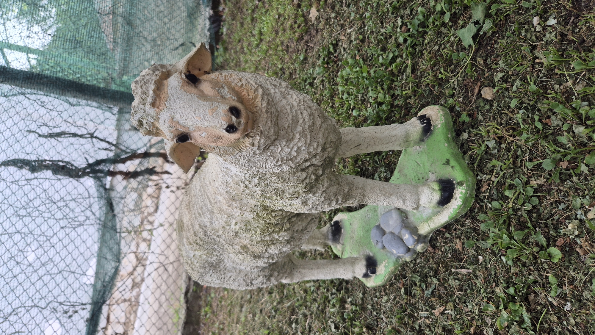 A concrete statue of a sheep stands on a green, irregular-shaped base with several gray stones on it. The sheep statue is white with some dirt and moss on it. It has black eyes and black hooves. The base is placed on the ground, which is covered with dry leaves and green grass. In the background, there is a green mesh fence. The scene is outdoors. The image was taken in the city of Doirentsi, Bulgaria.