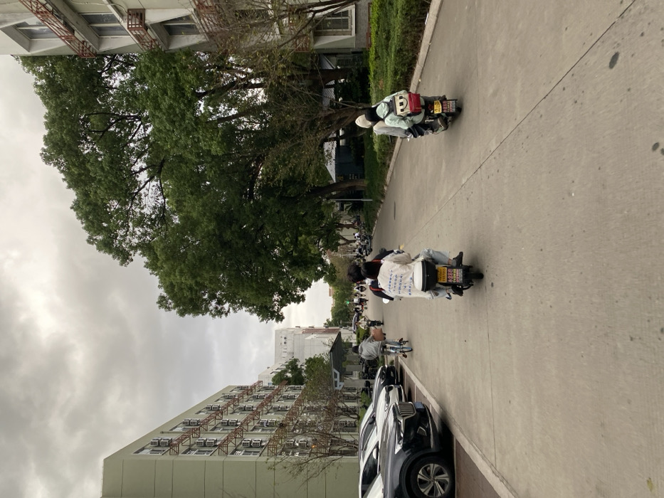 The image depicts an outdoor scene on an overcast day in what appears to be a campus or residential area within Yinzhou District, China. A grey, cloud-covered sky dominates the upper portion. The foreground features a concrete paved thoroughfare with several individuals and vehicles.
On the left side of the thoroughfare, a row of at least three parked passenger vehicles, including a dark SUV, a white SUV, and a black SUV, are visible. Adjacent to these vehicles stands a light-green, multi-story building (at least six floors visible) characterized by numerous window-mounted air conditioning units attached to external orange-colored metal support structures. Another lighter-colored building is partially visible further down this side.
On the right side of the thoroughfare, large, dense green trees line the path, partially obscuring a multi-story building with light-colored walls and external window railings. Landscaped green shrubbery is also present along this side.
In the immediate foreground, two electric scooters are moving away from the viewer. The scooter on the left carries two individuals; the driver wears a white long-sleeved shirt with dark script on the back and dark pants, while the passenger has dark hair. This scooter is equipped with a black storage box and displays a yellow license plate. To the right, a second electric scooter also carries two individuals; the passenger is wearing a light-colored hooded top and a light-green backpack featuring a prominent smiley face design. This scooter also has a yellow license plate.
Further down the thoroughfare, an individual is observed riding a blue bicycle. Multiple other pedestrians and individuals on electric scooters are visible in the mid-ground and background, suggesting active movement along the path. A green traffic light is discernible in the distance. The paved surface appears to be concrete.