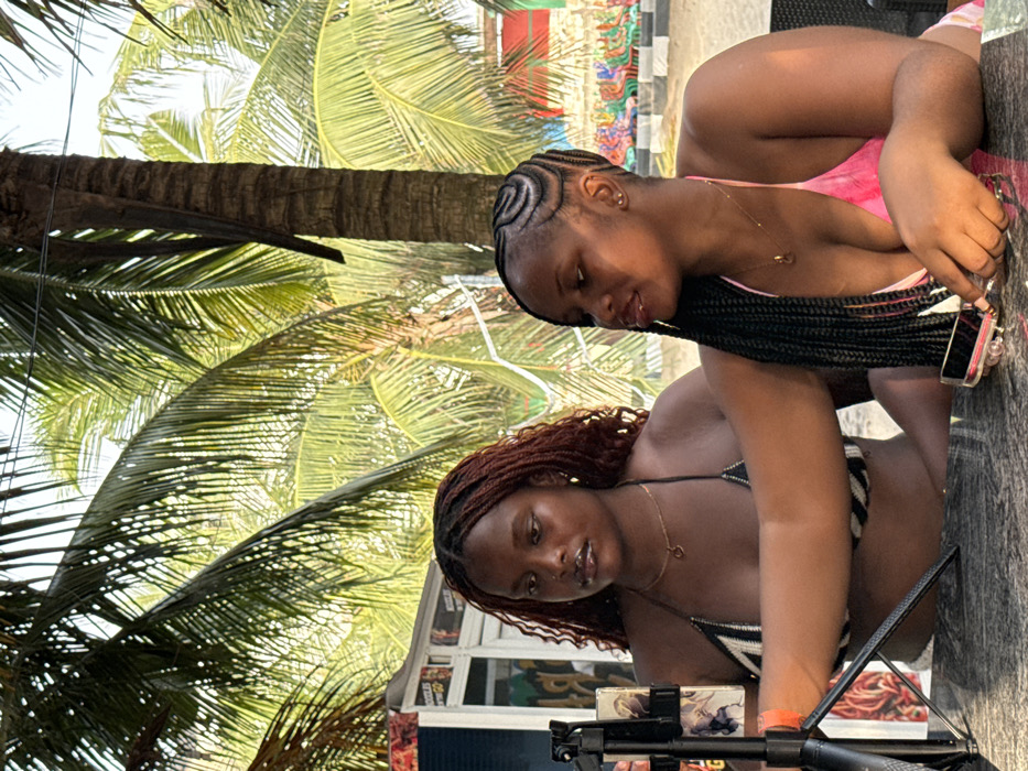 Two women in swimwear pose under a palm tree. The woman on top wears a pink tie-dye swimsuit, and her hair is styled in cornrows. The other woman wears a black and white striped bikini top and has wavy, auburn hair. Both wear necklaces. The woman in the bikini appears to be setting up a camera or phone on a tripod. The background includes palm trees and a building.