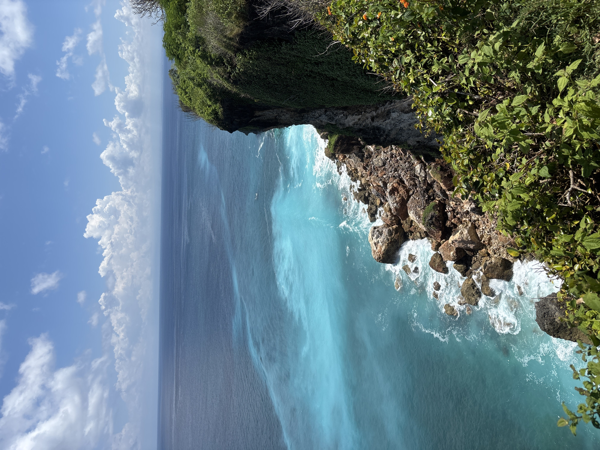 The image depicts a coastal scene in Uluwatu, Indonesia. A rocky cliff covered in dense green vegetation extends over the turquoise ocean. Waves crash against the rocks below. The sea transitions from a lighter, more vibrant blue near the shore to a darker, deeper blue further out. The sky, visible to the left, features a blue expanse with scattered white clouds.