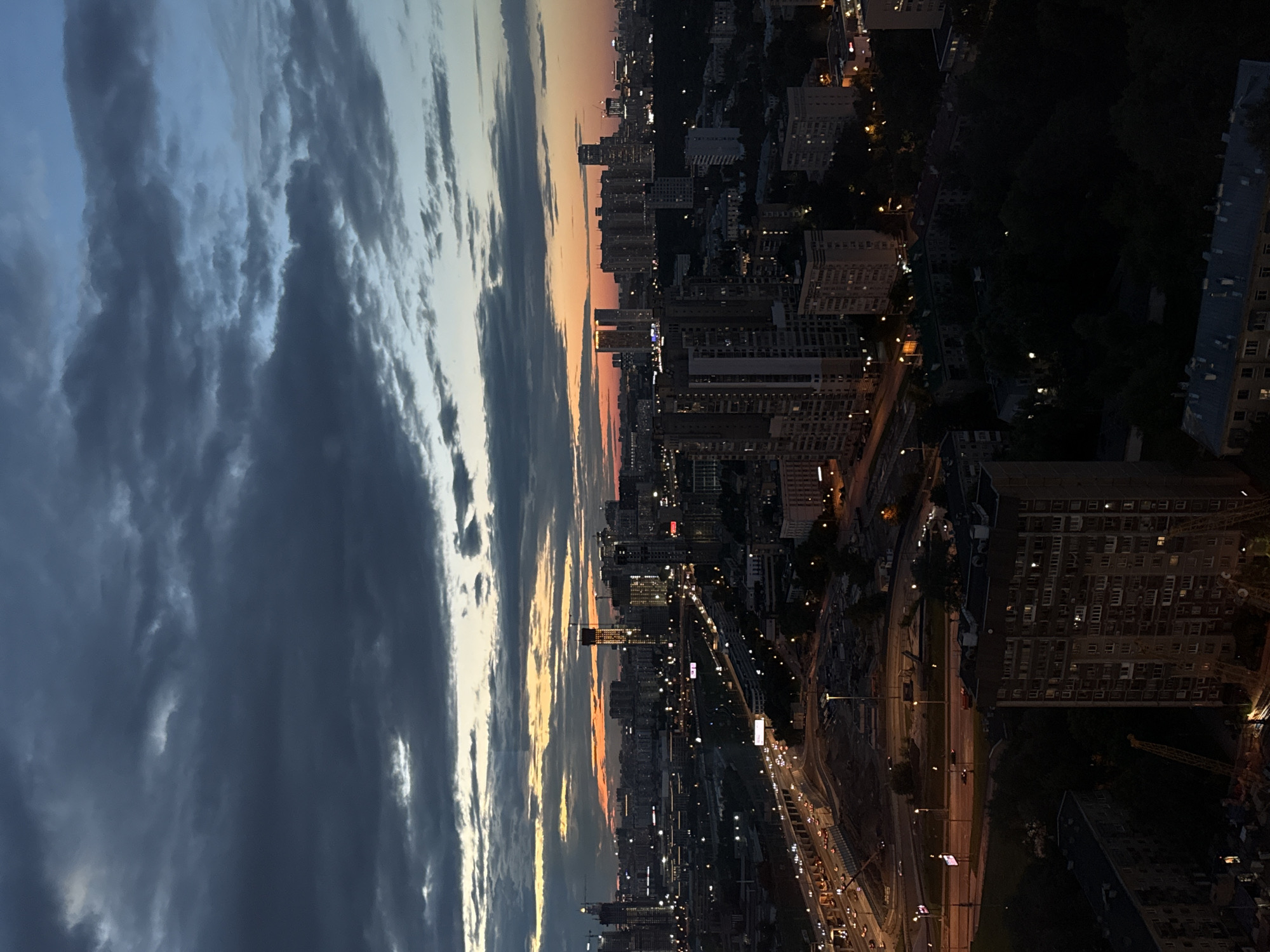 This is a high-angle, wide shot of a city skyline at dusk. The sky is a deep blue with heavy, dark clouds, illuminated by a strip of orange and yellow sunset near the horizon. Below the sky, a sprawling urban landscape is visible, characterized by numerous buildings of varying heights, including residential apartment blocks, office towers, and construction sites. Lights from buildings and street lamps are beginning to illuminate the city. A multi-lane highway with visible car traffic runs through the middle ground, appearing to be elevated or part of a complex interchange. The foreground is dominated by darker, lower-rise buildings and trees, with some construction cranes visible. The scene suggests a busy, developed metropolitan area. The location is identified as the city of Khimki, Russia. There are no visible people in the image.