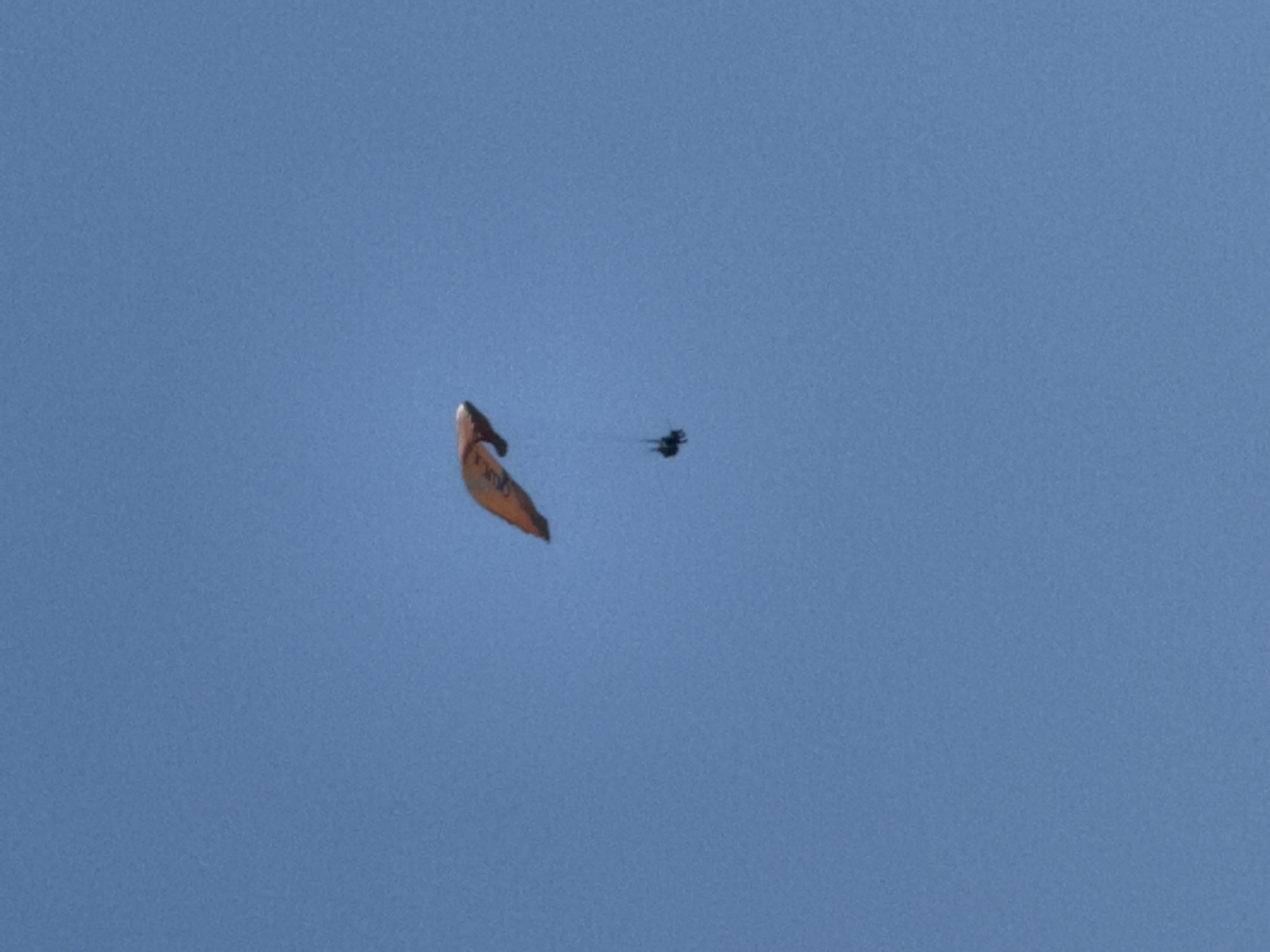 An orange and white paraglider is soaring through a clear blue sky over Boreti, Montenegro. The paraglider, with two people harnessed underneath, is in the upper center of the frame. The paraglider has some visible markings or logos on its surface. The image captures a moment of free flight, with no other objects or people visible in the frame. The vast expanse of the sky suggests an open area, likely a scenic spot in Boreti known for paragliding activities.