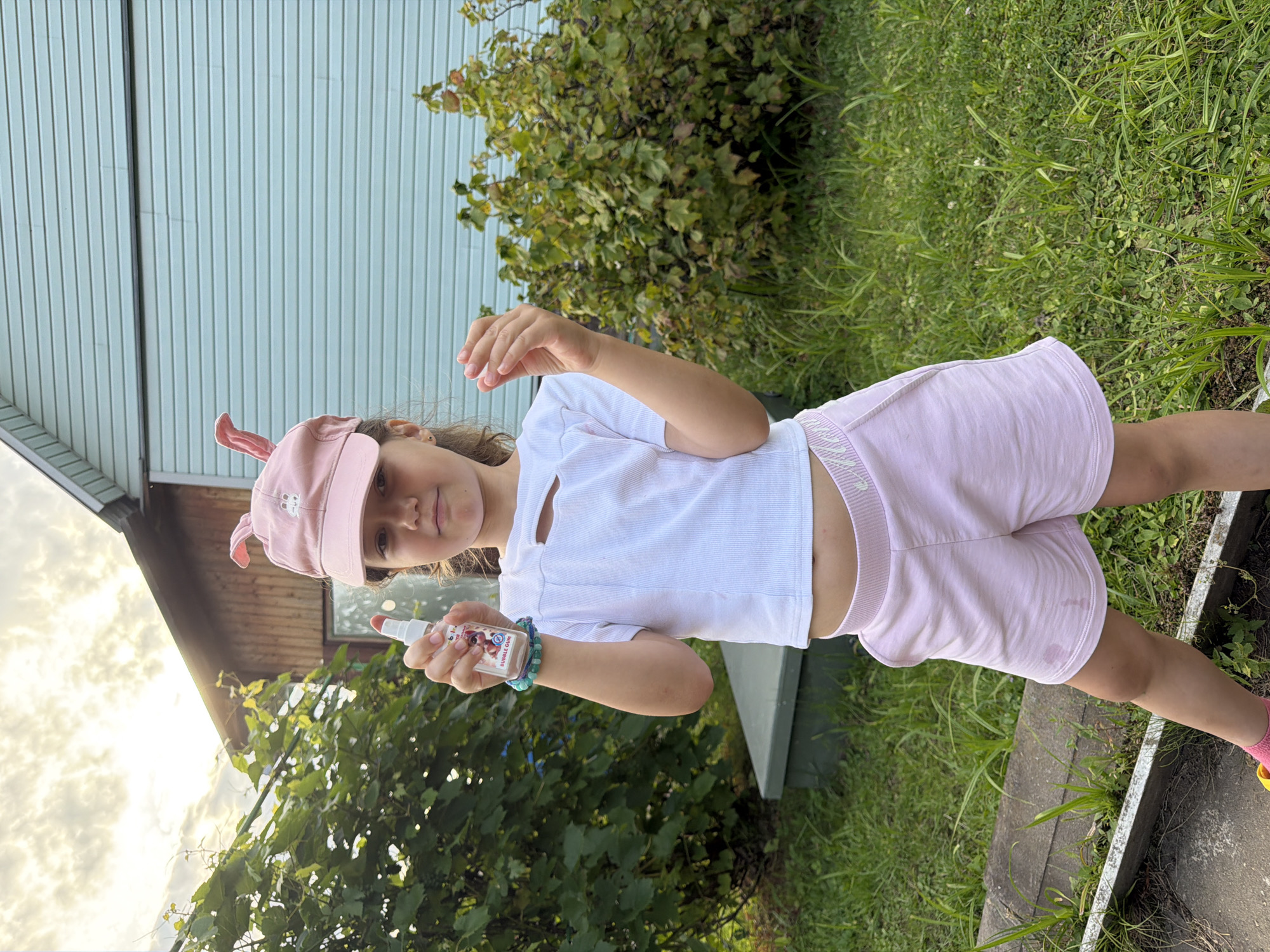 A young girl, wearing a pink baseball cap with bunny ears and a white t-shirt, stands outdoors in front of a light blue building with white vertical siding. She is holding a small bottle of what appears to be hand sanitizer. The bottle has a colorful label with text that includes "BUBBLE GUM". She is also wearing light pink shorts and a beaded bracelet. The setting is grassy with some greenery and foliage visible. The lighting suggests it could be late afternoon or early evening, with a cloudy sky visible in the background. The girl has a neutral expression. The location is identified as Nizinka SNT, Russia.