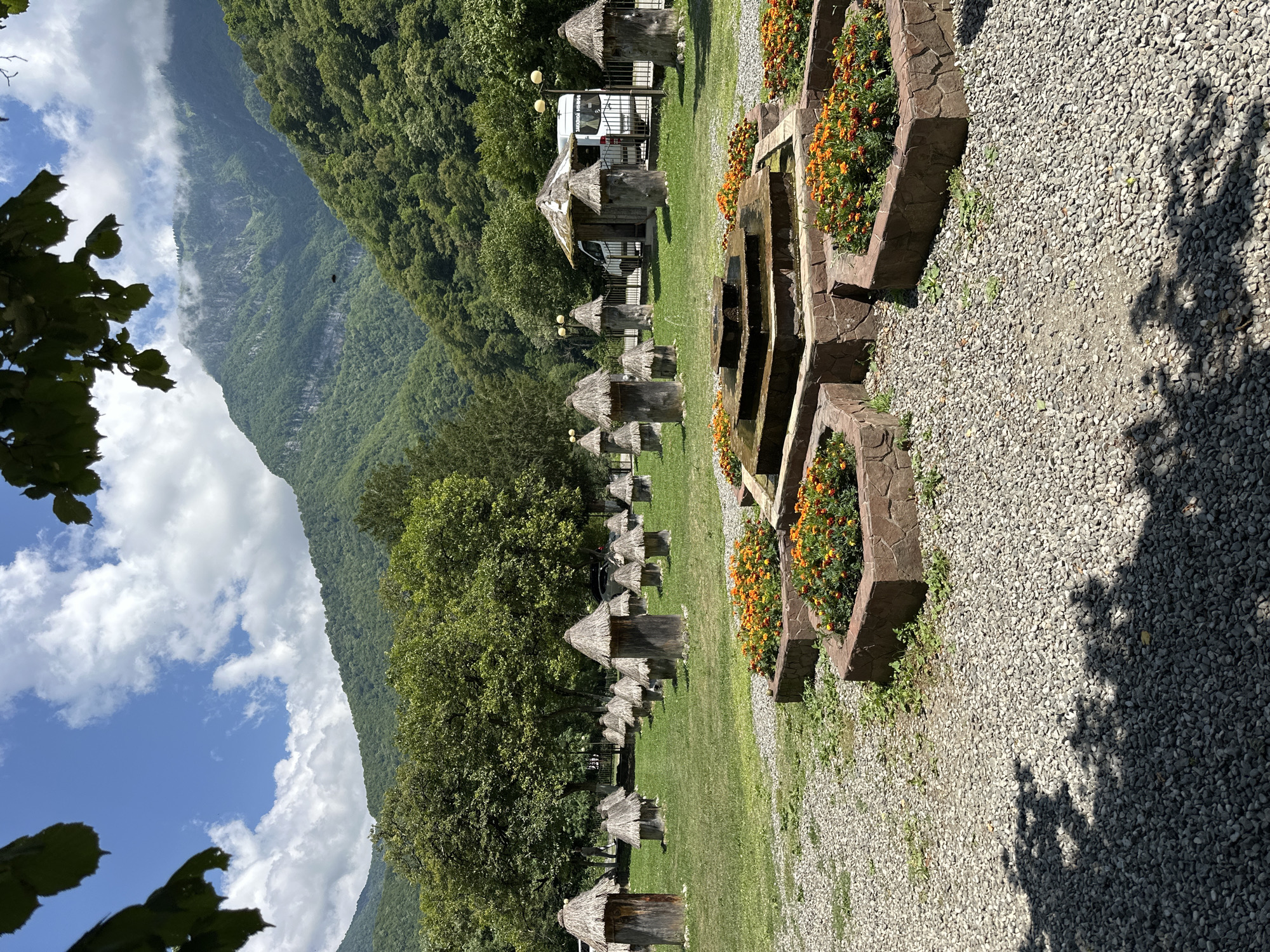 In the city of Bzypta, Abkhazia, a picturesque scene unfolds with a focus on a collection of traditional beehives. The beehives, approximately a dozen, are arranged in neat rows on a grassy field. Each hive is a hollowed-out log, topped with a conical thatched roof, giving them a rustic and charming appearance. In the foreground, a multi-tiered stone fountain with water flowing adds a soothing element to the environment. Surrounding the fountain are geometric-shaped flower beds filled with vibrant orange marigolds. Beyond the beehives and fountain, a dense forest of green trees blankets the lower slopes of majestic, mist-shrouded mountains. A white van and a small wooden structure are visible in the background, suggesting a visitor center or service area. The sky is a bright blue, dotted with fluffy white clouds, creating a serene and inviting atmosphere. The overall impression is one of natural beauty, traditional craftsmanship, and peaceful coexistence.