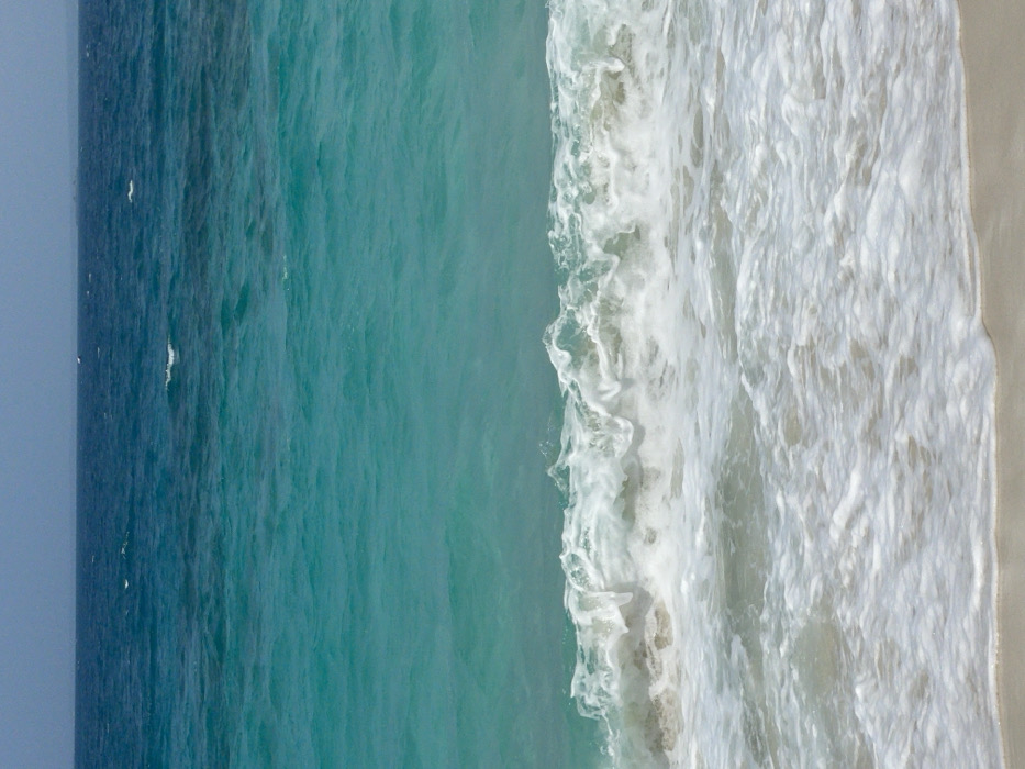 The image displays a coastal scene in Zanzibar, Tanzania, facing the open sea. A narrow strip of light-colored sand occupies the bottom fifth of the frame. Immediately above the sand, white foamy waves are actively breaking, indicating surf conditions. The water in the mid-ground is a clear, vibrant turquoise, exhibiting numerous ripples and small swells. Further out, the ocean transitions to a deeper blue, extending to a distant horizon line. The sky above the horizon is a hazy, light blue. Multiple small, dark, indistinct shapes are barely perceptible on the far oceanic horizon, consistent with distant vessels or very low-lying land. No individuals, prominent vegetation, or identifiable man-made structures are visible.