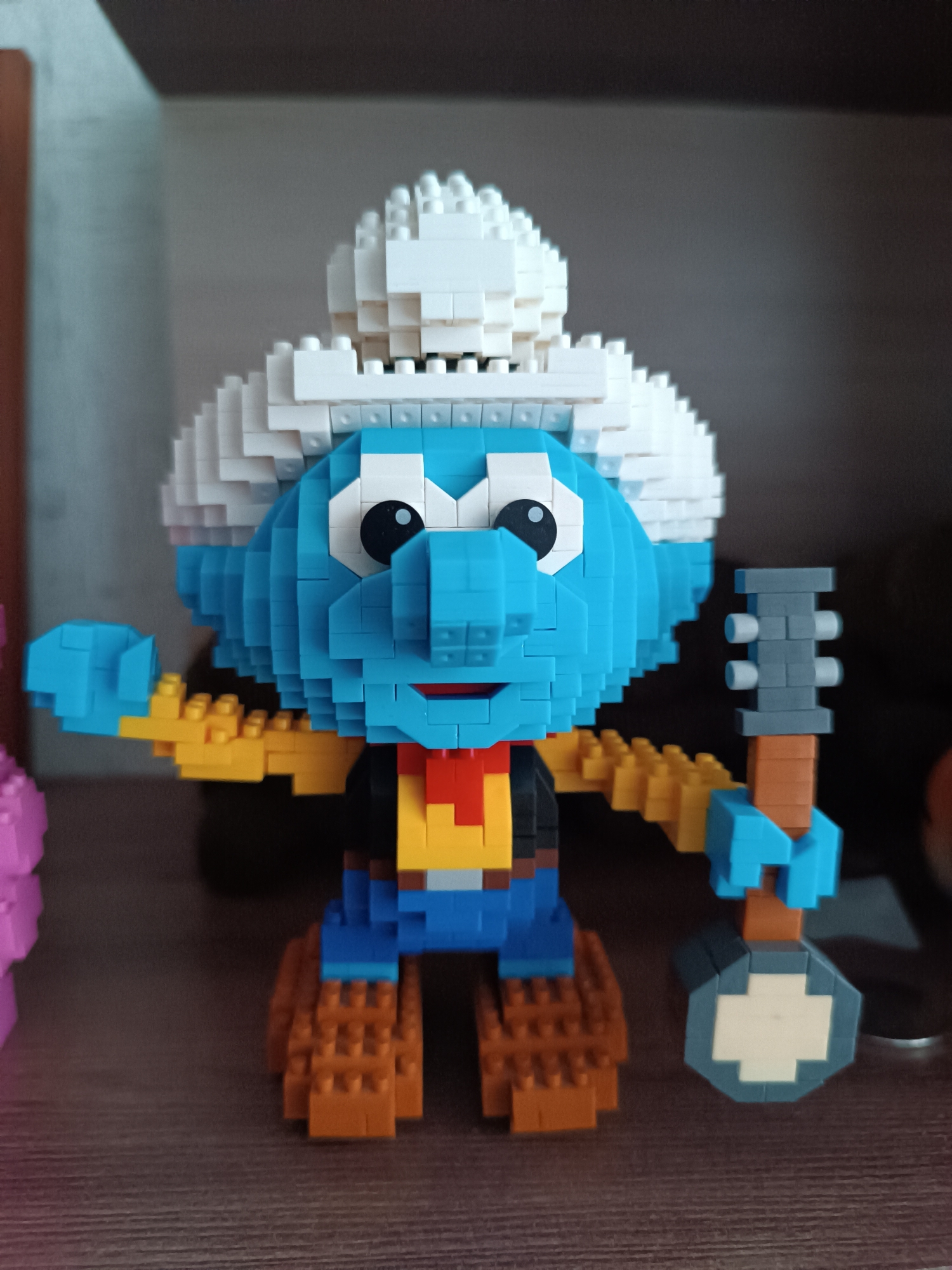 A front-facing, medium close-up shot captures a blue Smurf character constructed from small, interlocking building blocks, resembling a pixelated figurine. The Smurf is depicted as a cowboy, wearing a large white cowboy hat, a yellow shirt with a red bandana around its neck, blue pants, and brown boots. Its face is blue with large, white eyes featuring black pupils, and a small, red, smiling mouth. The figure holds a banjo-like instrument in its right hand, which has a brown neck, grey tuning pegs, and a grey and off-white circular body. The figure stands on a dark brown, wood-grained surface, possibly a shelf or table. In the blurry background, a light grey wall is visible behind the figure, with a darker horizontal surface (another shelf or the underside of one) above. A hint of another pink block figure is partially visible on the far left of the frame. The lighting suggests an indoor setting. No visible text is present.
