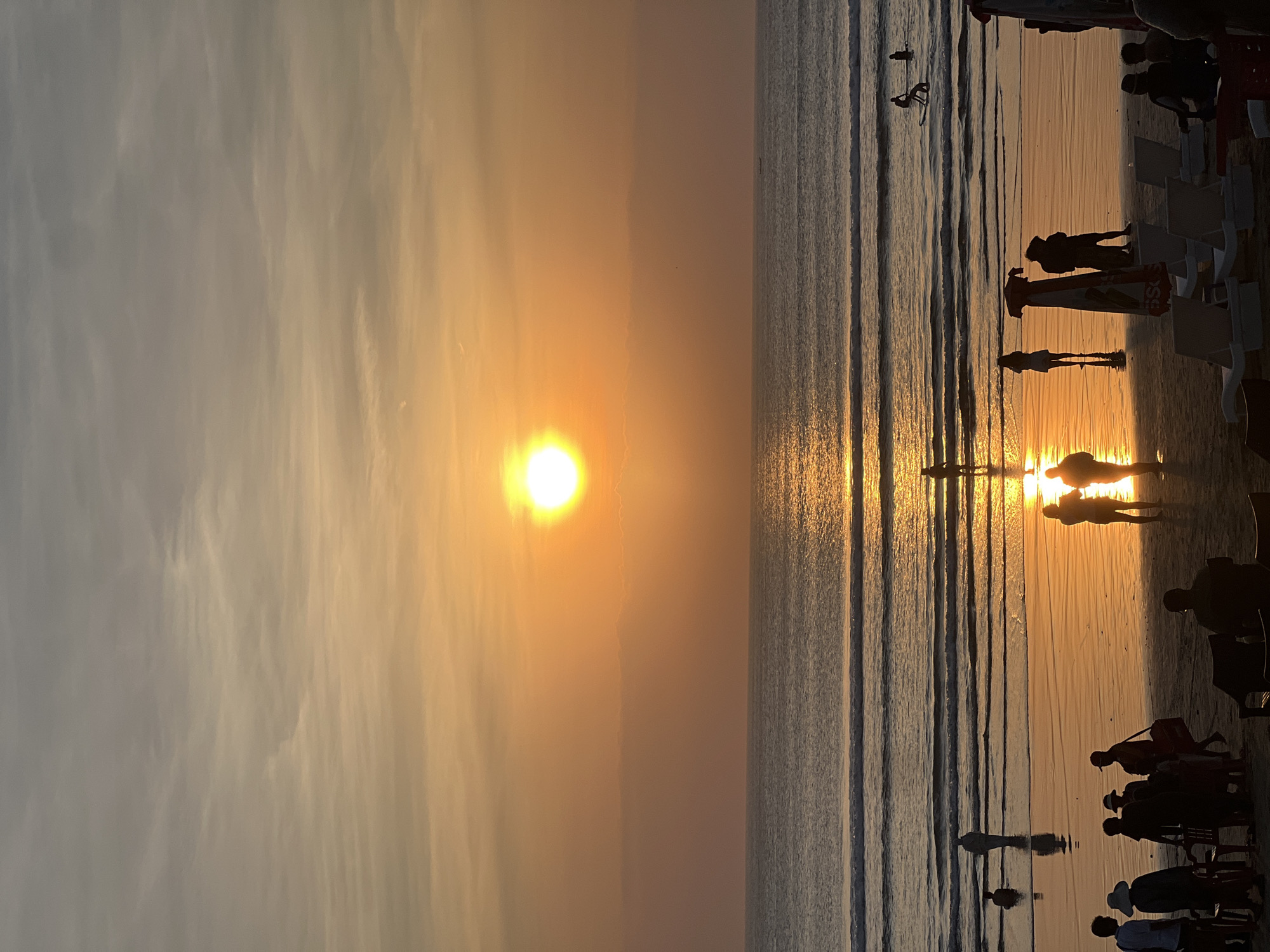 A vertical, eye-level shot captures a vibrant sunset over the ocean at Kuta Beach, Indonesia. The sun, a bright orb of light, is positioned high in the sky, casting a warm, golden glow across the scene. The sky is a gradient of soft oranges and grays, with wispy clouds scattered throughout. The ocean is a deep blue, with gentle waves rolling towards the shore. The wet sand on the beach reflects the sunset, creating a mirror-like effect.
In the foreground, silhouetted figures of people are scattered across the beach. Some are standing and conversing, while others are seated on beach chairs, presumably enjoying the sunset. A few individuals are seen walking along the shoreline, their figures elongated by the low angle of the sun. In the middle ground, more people are visible, some wading in the shallow water, while others are further out, with a few appearing to be surfing or engaged in watersports. The reflections of the people and the sun shimmer on the water's surface. The overall atmosphere is serene and picturesque, with the warm hues of the sunset creating a sense of tranquility.