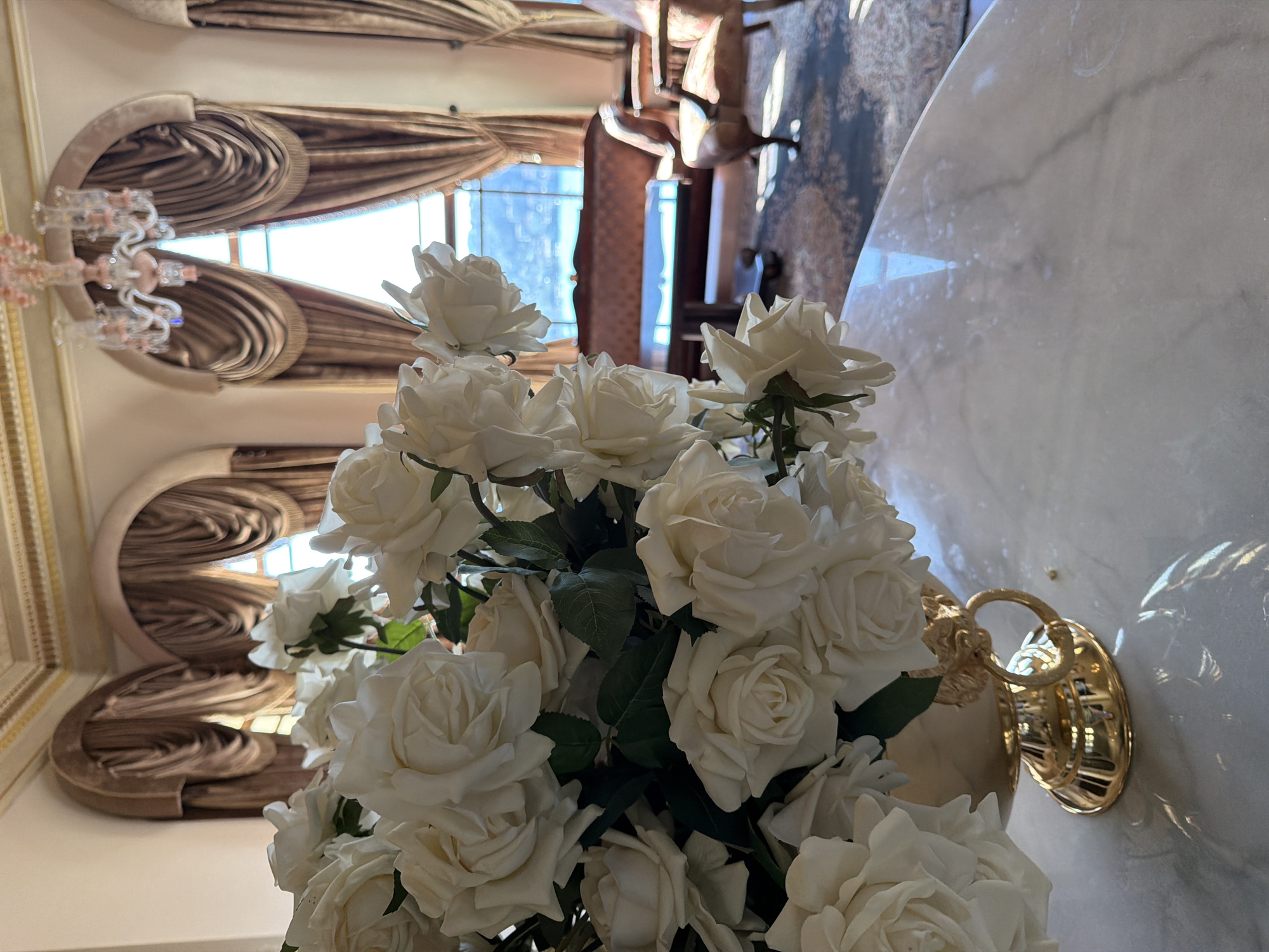A bouquet of white roses in a gold vase sits on a marble table in a sunlit room with large arched windows draped with heavy brown curtains. In the background, a view of the Bosphorus strait in Üsküdar, Turkey, is visible through the windows, with a brown sofa and armchair placed on an oriental rug. A chandelier hangs from the ornately decorated ceiling.