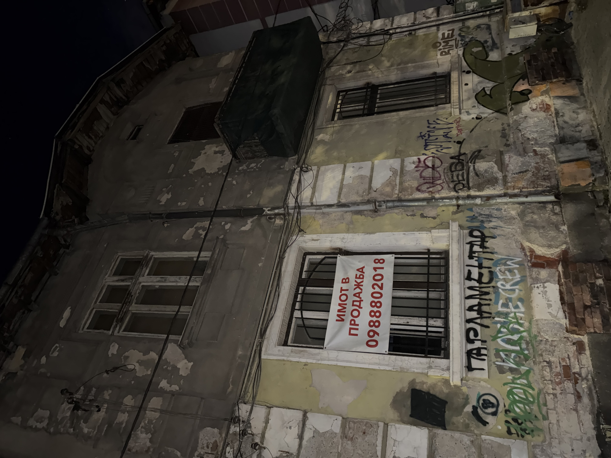 The image captures a dilapidated, multi-story building at night in Sofia, Bulgaria. The building's facade is heavily weathered, with large sections of paint and plaster peeling off, revealing the underlying structure and some exposed brickwork. The dominant colors are muted grays, yellows, and the dark tones of the night sky.
Several windows are visible on the building. On the lower level, a window with security bars features a white banner with red Cyrillic text. The banner clearly states "ИМОТ В ПРОДАЖБА," which translates to "PROPERTY FOR SALE," followed by a phone number: "0988802018." Above this, another window also has security bars. On the upper left, a darker, multi-paned window appears to be either boarded up or deeply recessed.
Numerous electrical wires run haphazardly across the building's exterior, adding to the unkempt appearance. An outdoor unit, possibly an air conditioner, covered in dark material, hangs precariously on the upper section of the building. Extensive graffiti covers various parts of the walls, indicating an urban setting. Visible graffiti includes "RIME" in green, "ДЕДА" (DEDA), and "ПАРАЛАМЕНТАР" (PARALAMENTAR), with "CREW" written below it. The low light of night emphasizes the shadows and worn textures of the building. No people are visible in the frame.