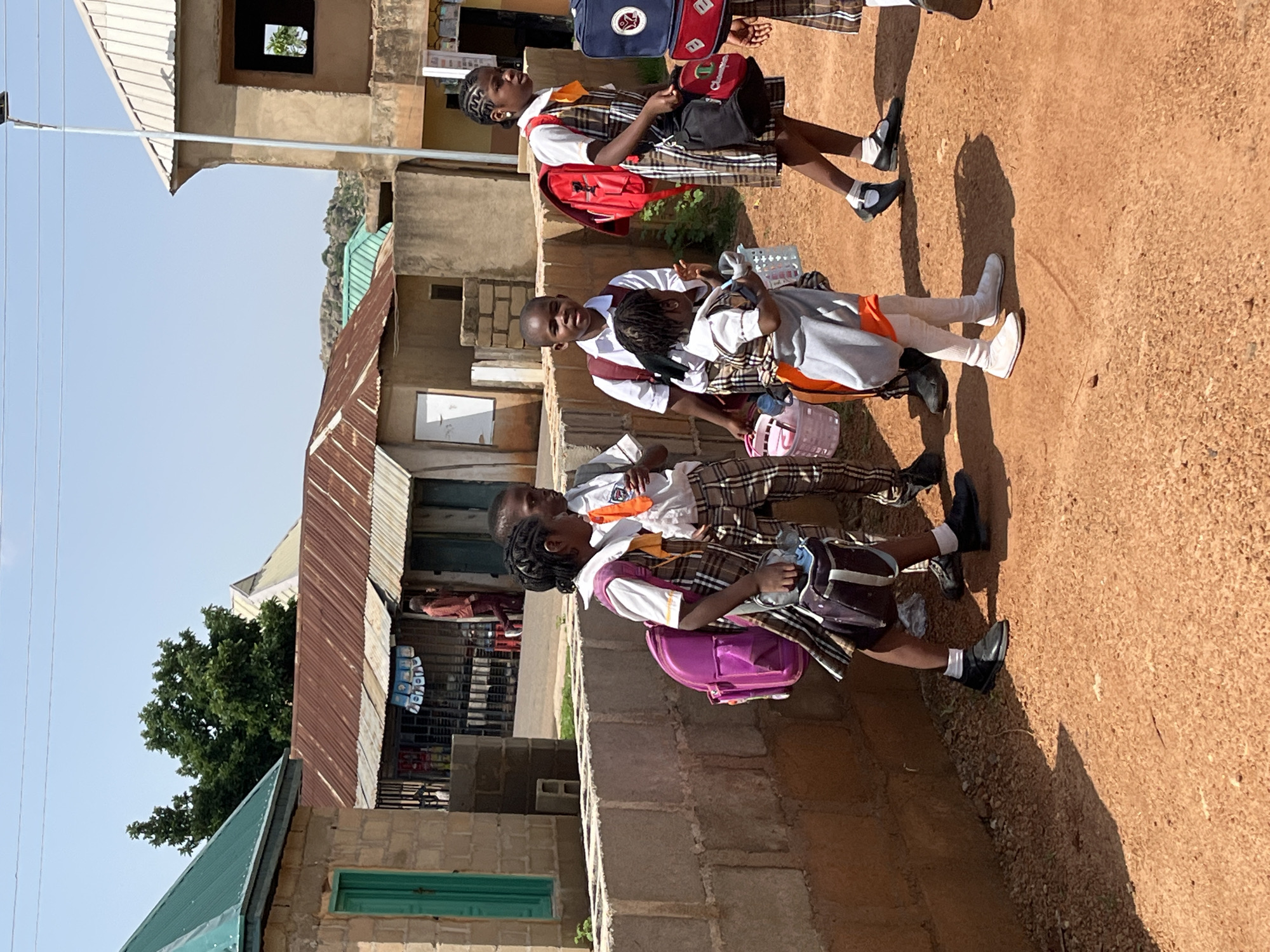 In Barakin Akawo, Nigeria, on a sunny day, a group of four children, dressed in school uniforms, stand on a dirt path near a brick wall. The uniforms consist of plaid shirts and trousers or skirts, worn over white collared shirts. The children are carrying backpacks, lunchboxes, and other school supplies. The youngest child, a girl in the center, wears a grey sweater over her uniform and holds a white plastic basket. To her left, a girl with braided hair wears a pink backpack and a dark-colored satchel. To her right, a boy with short hair is dressed in a plaid vest over his uniform, carrying a red backpack. Another child, mostly out of frame to the right, also carries a red backpack. In the background, there are buildings with corrugated metal roofs, a street, and trees. A person in a reddish-brown outfit walks in the distance.