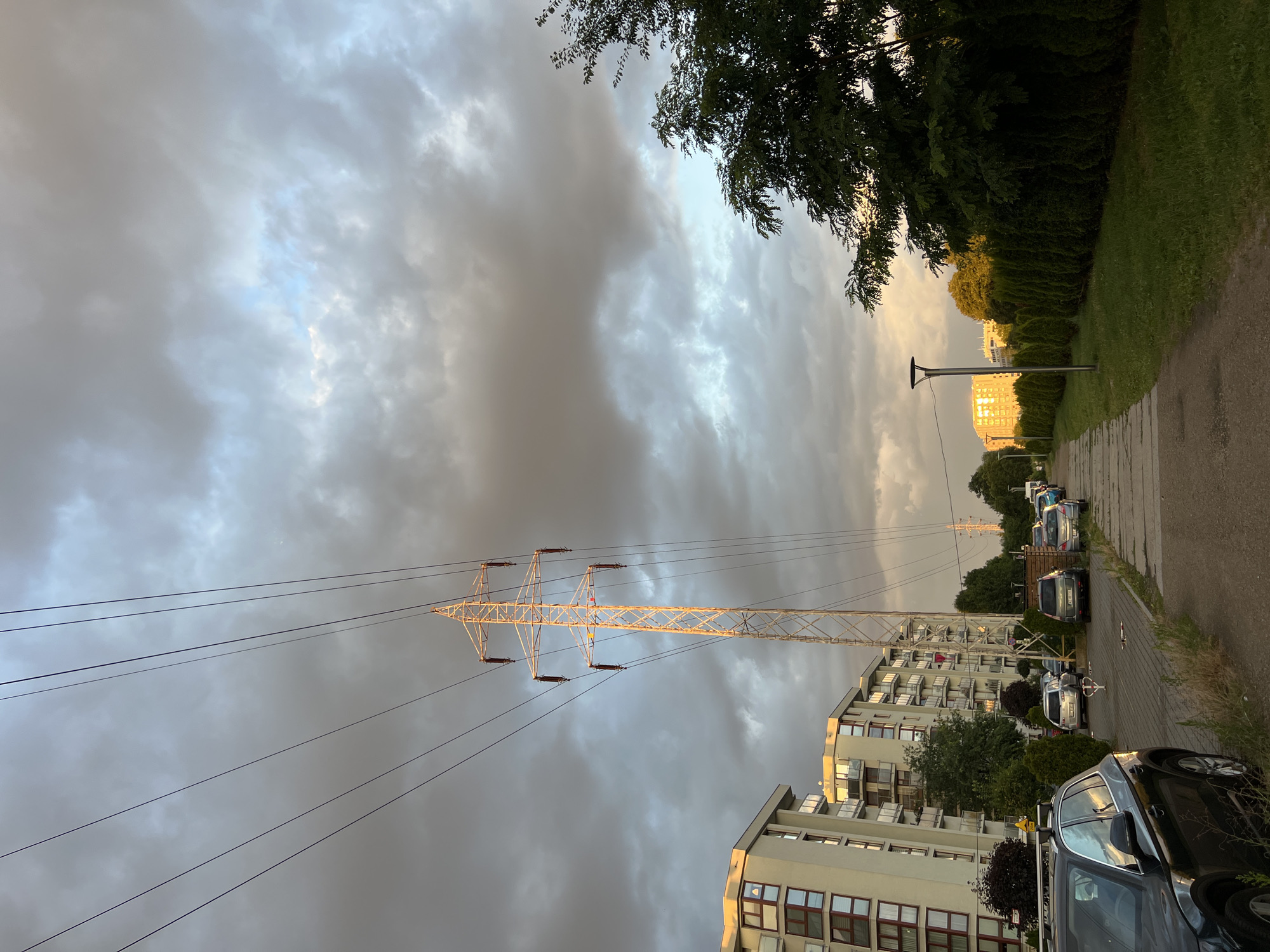 The image depicts a street in Krakow, Poland, with a power line tower dominating the sky. The sky is filled with dramatic, overcast clouds, with patches of sunlight breaking through, casting a warm glow on the buildings. On the left, modern apartment buildings with balconies line the street, reflecting the golden light. A row of parked cars is visible along the side of the road. A paved walkway runs alongside a well-maintained hedge, with green grass on the other side. In the distance, more buildings are visible, illuminated by the setting sun. The scene appears to be a residential area, with the power line tower suggesting urban infrastructure.