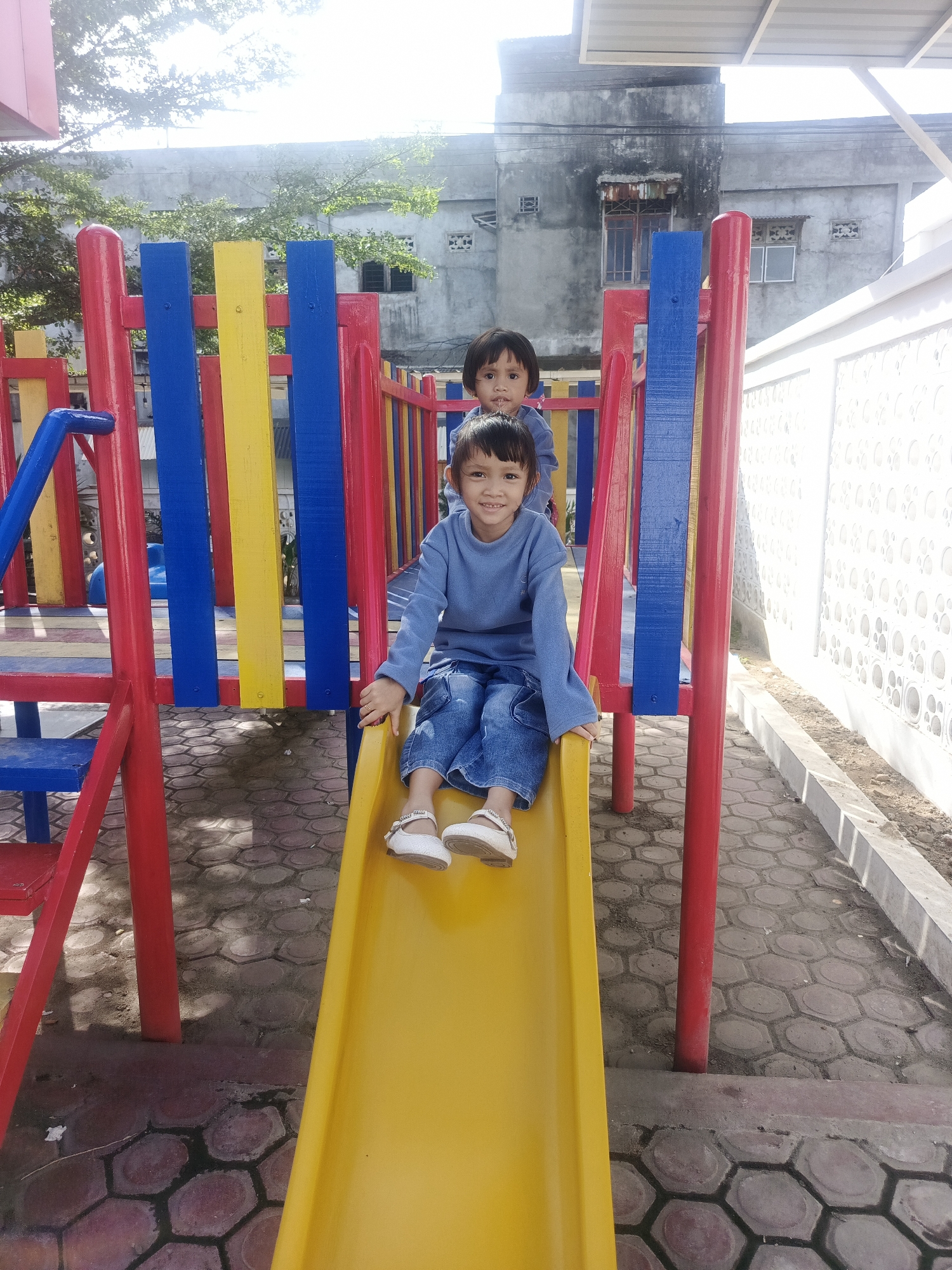 The image displays an outdoor scene in Pantegajah, Indonesia, featuring two young children on a multi-colored playground structure under bright daylight. In the foreground, a child, likely of East Asian descent, is seated on a yellow slide, facing the camera with a smile. This child is wearing a light blue long-sleeved shirt, blue jeans, and white flat shoes with straps. Behind and slightly above, a second child, similarly attired in a light blue long-sleeved shirt and blue jeans, is partially visible from the chest up, positioned further back on the playground structure and looking towards the camera.
The playground equipment consists of a yellow slide, red structural supports, blue steps, and vertical barriers made of alternating blue and yellow planks. The ground is paved with hexagonal concrete blocks, exhibiting variations in color from grey to reddish-brown, with some darker, potentially damp or shaded areas. In the background, a weathered, multi-story grey concrete building with visible windows and signs of discoloration or rust is present, partially obscured by sparse green foliage. To the right of the playground, a white wall with a repeating circular pattern in relief extends, alongside a segment of a white metal awning structure.