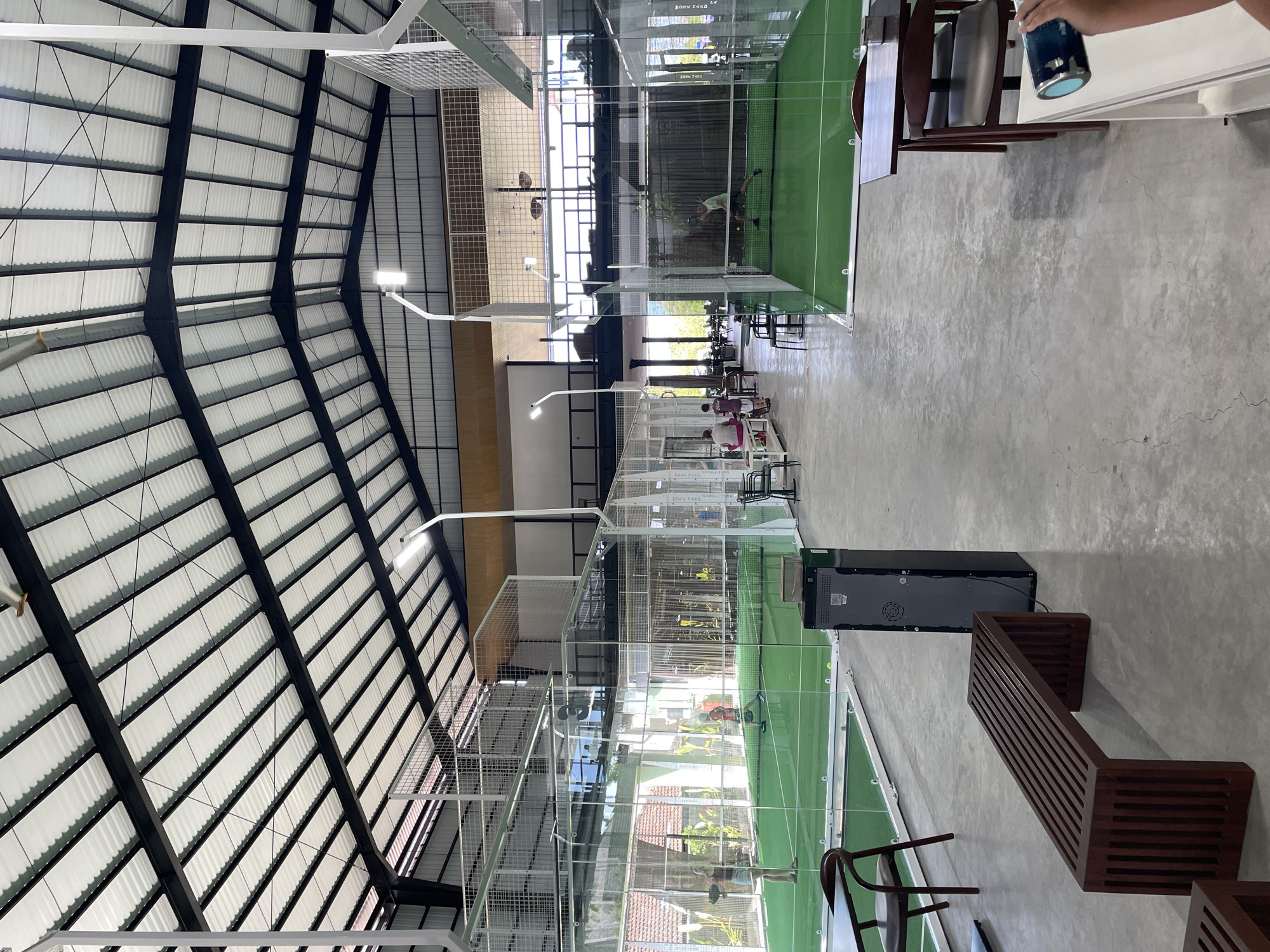 The image depicts the indoor paddle tennis courts at an establishment in Kuta Selatan, Indonesia. The scene features multiple enclosed paddle tennis courts with green turf flooring and glass walls. Several individuals are present, some actively playing paddle tennis, while others are seated or standing around the courts. In the foreground, a dark wooden bench and a dark grey equipment cabinet are visible on the concrete floor. Tables and chairs are set up in a seating area to the right of the frame. The facility has a high, industrial-style ceiling with exposed beams and white corrugated roofing, and is illuminated by artificial lighting. Sunlight filters in from the open sides and large windows, indicating a daytime setting. The overall environment suggests a sports and leisure complex.