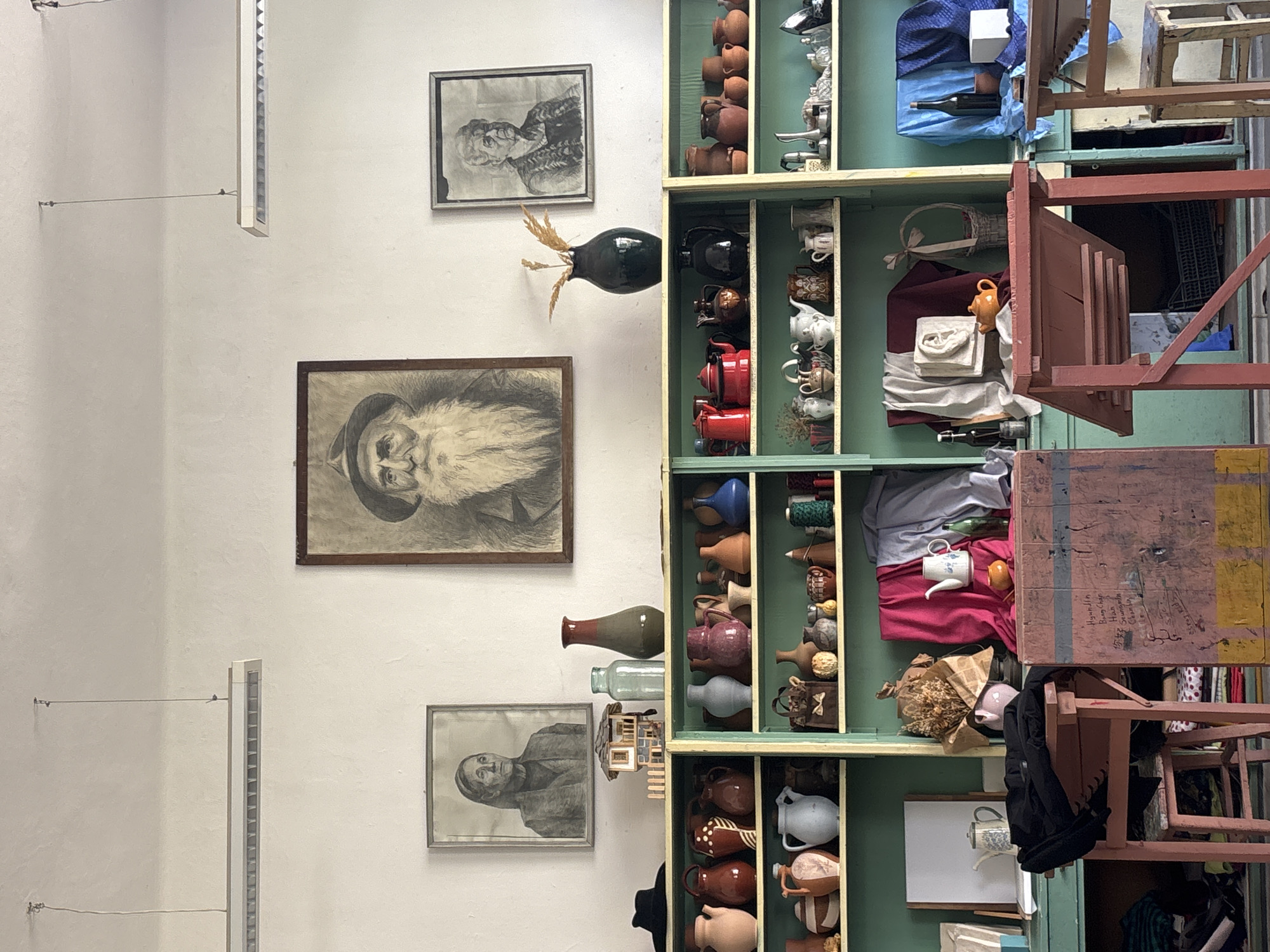 The image captures an interior wall in Troyan, Bulgaria. On the wall, three framed portraits hang vertically, two of older women and one of an older man with a beard. A dark-colored vase containing dry foliage is positioned between the upper two portraits. A light fixture is mounted vertically on the left side of the wall. To the right of the portraits, a tall, narrow, multi-tiered shelving unit, painted green, displays an eclectic assortment of objects: pottery, teapots, kitchenware, textiles, and a few glass bottles. A series of chairs, some stacked, are partially visible to the right of the shelving unit.