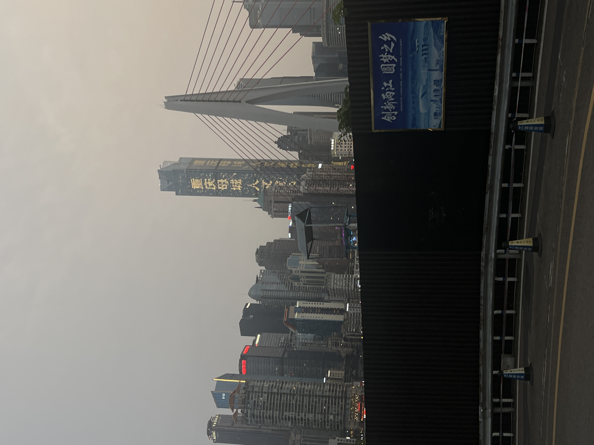 The image captures a wide shot of a city skyline at dusk, with a focus on a modern bridge and a cluster of skyscrapers. The sky is overcast and hazy, casting a muted light over the scene. In the foreground, a dark, ribbed fence runs horizontally across the image, partially obscuring the view of the city. A road with yellow and white lane markings is visible in the lower portion of the frame, with two traffic cones placed near the fence.
Dominating the center-right is a distinctive cable-stayed bridge with a single, large white pylon. The cables extend outwards, connecting to the bridge deck. To the left of the bridge, a tall, slender skyscraper is illuminated with vertical lines of yellow text. The text appears to be Chinese characters. Behind this prominent building are numerous other skyscrapers of varying heights and architectural styles, some with illuminated windows and signs. One building towards the far left has "ADBC" visible on its upper section.
On the fence in the lower right quadrant, a large blue and gold framed poster is affixed. The poster displays an artistic rendering of a cityscape with buildings and a body of water, along with Chinese text at the top. The location is identified as 江北城街道 (Jiangbei Cheng Subdistrict), China. The overall impression is one of urban development and modern infrastructure in a coastal or riverside setting, seen through a hazy atmosphere.