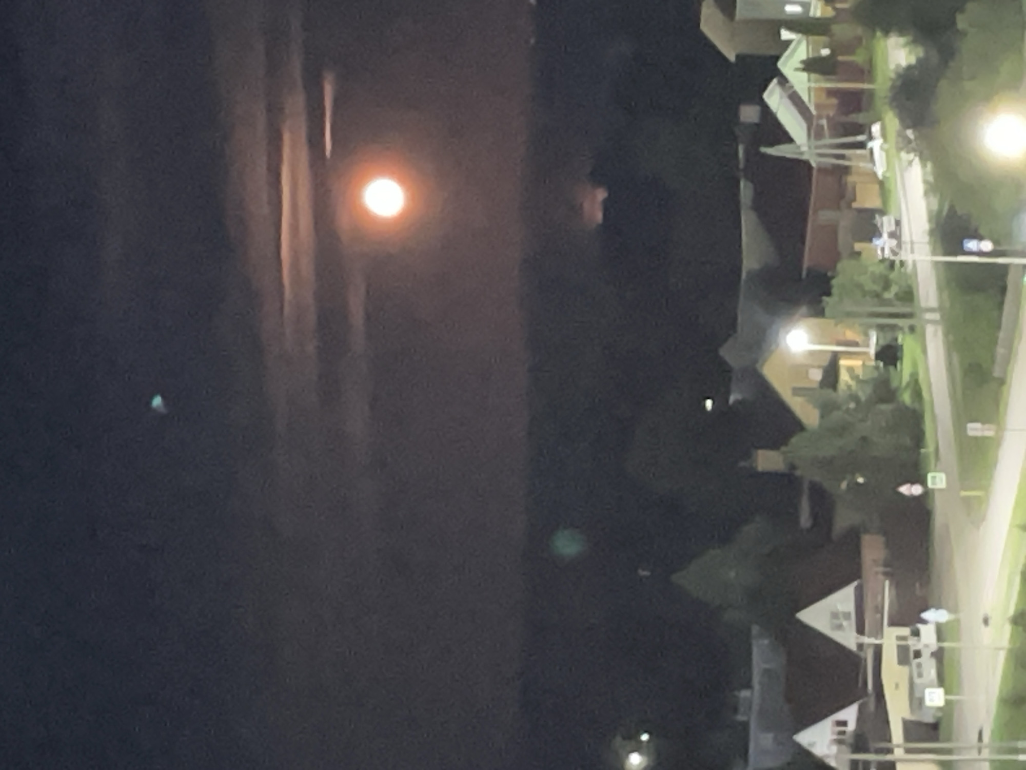 The image depicts a nighttime aerial view of a residential area in Laishevo, Russia, under a full moon. The moon, a vibrant orange sphere, is partially obscured by thin clouds, casting a soft glow over the landscape. Below, a cluster of houses with illuminated windows and dark rooftops is visible, nestled amongst trees and a winding road. A street light illuminates a section of the road, and several parked vehicles are scattered throughout the scene. The overall atmosphere is serene and quiet, characteristic of a peaceful evening in a suburban setting. In the sky, a faint, teal-colored object is visible to the left of the moon.