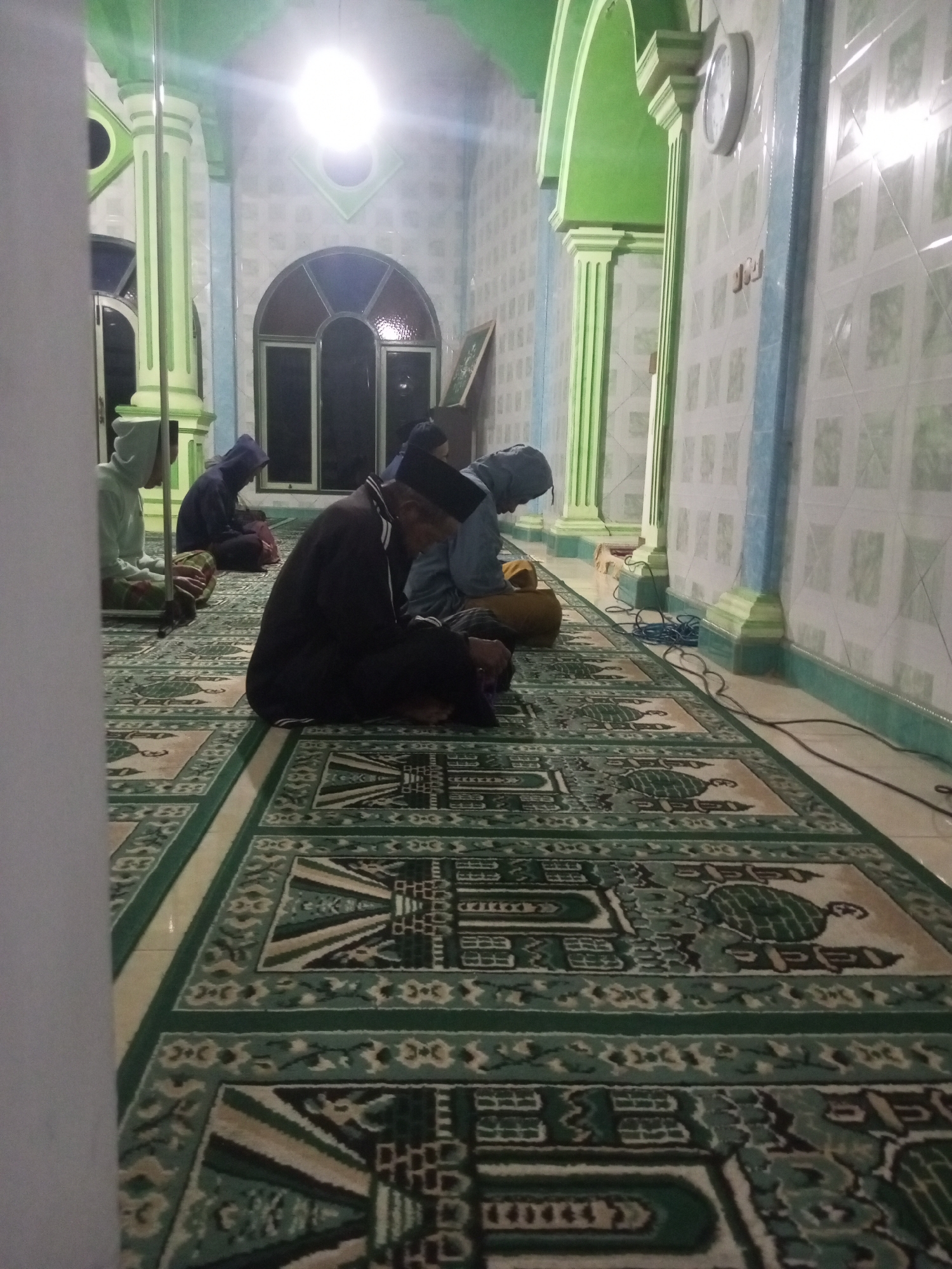 The image depicts a group of people praying inside a mosque. The mosque has a distinctive green and white color scheme, with green arches and columns. The floor is covered with a patterned carpet, predominantly in green and beige. Several individuals are seen sitting on the carpet in prayer positions. One elderly man in the foreground is wearing a black robe and a white skullcap, facing away from the camera. To his left, a younger person in a light blue hoodie is also praying. Further back, other individuals in similar attire are visible. The lighting appears to be artificial, likely from overhead lamps, casting a bright glow in some areas. The walls are tiled with a light green pattern. The overall impression is one of quiet devotion within a religious space. This scene is located in the city of Andongsili, Indonesia.