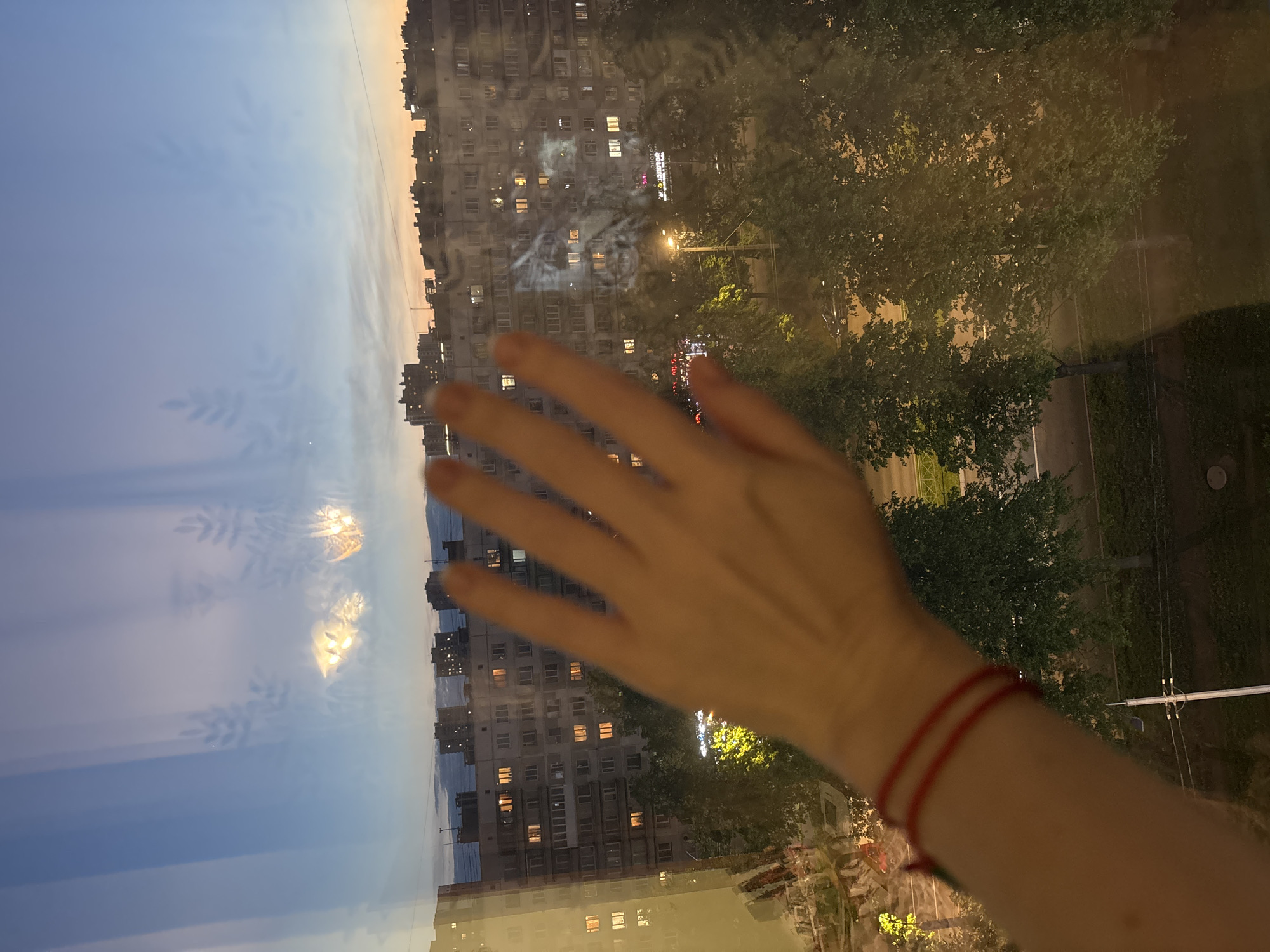 A person's hand is held up against a window reflecting a sunset sky, a high-rise apartment building with lit windows, and green trees. The hand has a red string bracelet on the wrist. The location is Vsevolozhsk, Russia, indicated by the typical architecture and urban setting.