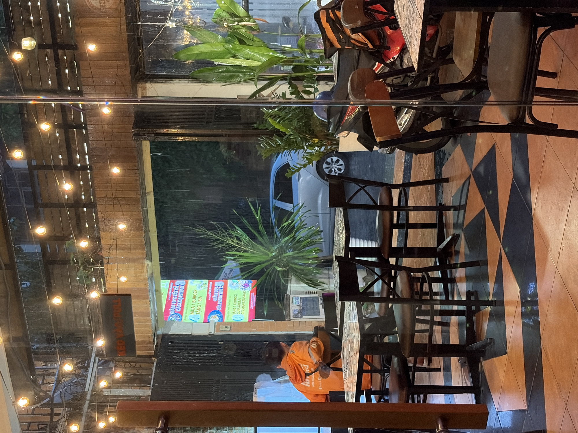 This is a medium shot of an outdoor seating area of a restaurant, possibly a fast-food establishment, on a rainy day in Thu Duc City, Vietnam. The foreground features several tables and chairs made of dark metal with wooden seats and backs. The tables have patterned tops, and the floor is tiled in a diamond pattern of orange and black.
In the midground, through a glass partition, a person wearing an orange hooded jacket is seated at a table, facing away from the camera. Behind them, a light blue car is parked outside. Various potted plants are visible, including a tall, spiky palm-like plant. A motorbike with delivery bags is parked to the right.
The background shows the entrance to the establishment, with a sign that reads "KEO VÀO PULL". Strings of fairy lights are suspended above the seating area, and the rainy weather is evident from the streaks on the glass and the wet appearance of the outdoor environment. The overall lighting is dim, with the fairy lights providing a warm glow.