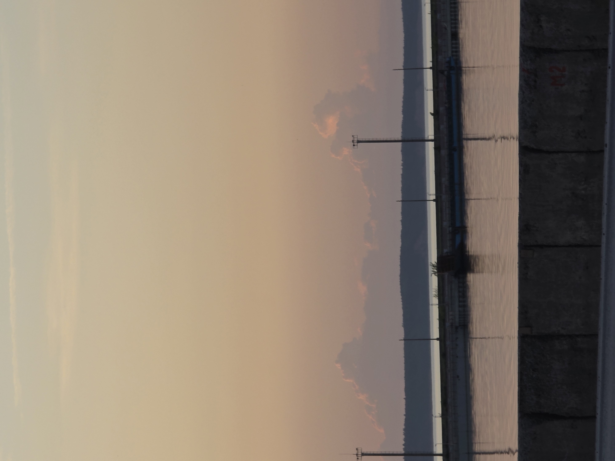 The image depicts a landscape near the city of Chaikovsky, Russia, likely at dusk. A calm body of water, possibly a river or lake, is visible on the right, reflecting the sky's colors. A concrete structure with numbered markings (possibly "M2") borders the water on the right side. A line of utility poles stretches along the water's edge, with the distant opposite shore barely visible. The sky transitions from a light orange/yellow near the horizon to a deeper pink/grey further up. There are no people visible. The overall atmosphere is still and serene.
