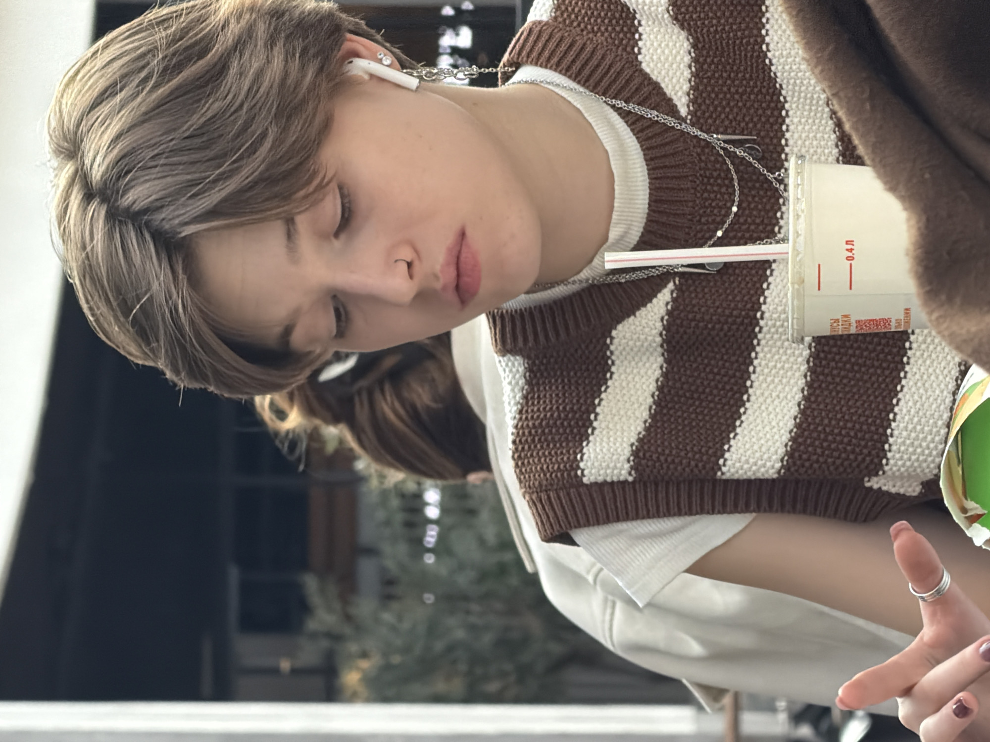 The original image presents a rotated close-up of a young individual in a relaxed setting, viewed from a slightly high angle. The image is oriented vertically, with the person's head towards the left side of the frame.
The central subject is a young person, likely a teenager or young adult, with short to medium-length, light brown hair styled with a middle part. Their eyes are closed or cast downwards, and their head is tilted to the side, conveying a sense of calm, contemplation, or rest. They have fair skin and their lips are slightly parted.
They are wearing white wireless earbuds in both ears, which are connected by a delicate silver chain that loops around their neck. A small silver stud earring is visible in their right ear. Their attire consists of a brown and white striped knitted vest layered over a white long-sleeved top, possibly a high-neck or turtleneck.
A white paper cup with a red and white striped straw is prominently featured in the foreground, positioned to the right of the person's face. The cup displays red Cyrillic text, including "0.4 Л" (indicating 0.4 liters), and smaller words such as "ВКУСЫ" (Tastes) and "НАПИТКИ" (Drinks), along with a QR code, strongly suggesting it contains a beverage from a cafe or fast-food establishment in Russia, consistent with the Saint Petersburg context.
In the bottom right corner, one of the person's hands is visible, featuring manicured nails painted with dark polish and two silver rings stacked on a single finger. The background is softly blurred, showing indistinct shapes and tones of dark grey and green, possibly indicating an outdoor view with foliage or blurred indoor elements. Another person's blurred head and hair can be faintly seen in the deeper background. The scene suggests a daytime moment of quiet relaxation or focused attention.