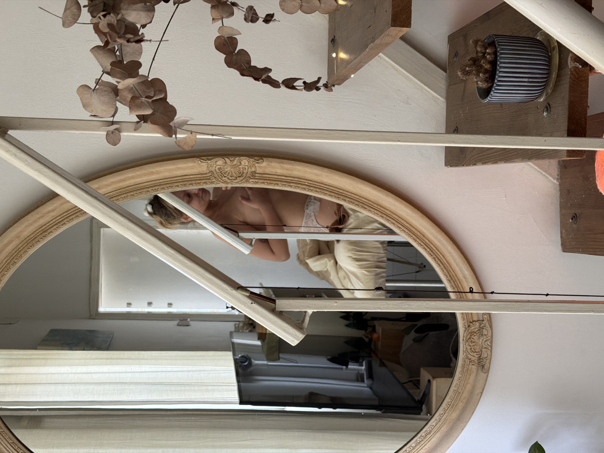 The scene depicts an interior view reflected in an oval mirror with a decorative, light-colored frame. The reflection shows a woman wearing a white bra and a light-colored robe or blanket. Her reflection also includes a window with white blinds and a television. Outside of the mirror, there is a plant with dried leaves hanging from the top, a wooden shelf with a small blue and white striped pot, and a white shelf. The presence of Hebrew letters on the television and on objects within the reflection indicate the location might be in Israel. However, without further evidence, it is not possible to definitively pinpoint the location as Tel-Aviv.