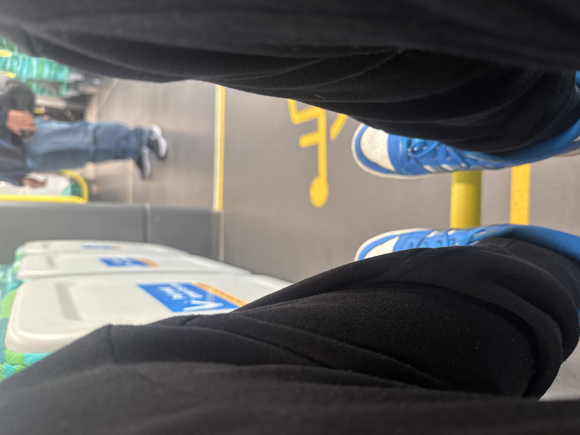 The image appears to be a first-person perspective shot, focusing on the legs and shoes of the person taking the photograph. The person is wearing black clothing, likely pants or leggings, and blue and white sneakers. The background is blurry but shows the inside of a building, possibly a shopping mall or public space in Bateman, Australia. A person wearing blue jeans and grey shoes is laying on a bench. There are rows of white boxes with blue labels, suggesting this could be a retail or storage area. A yellow musical note is visible on the floor.