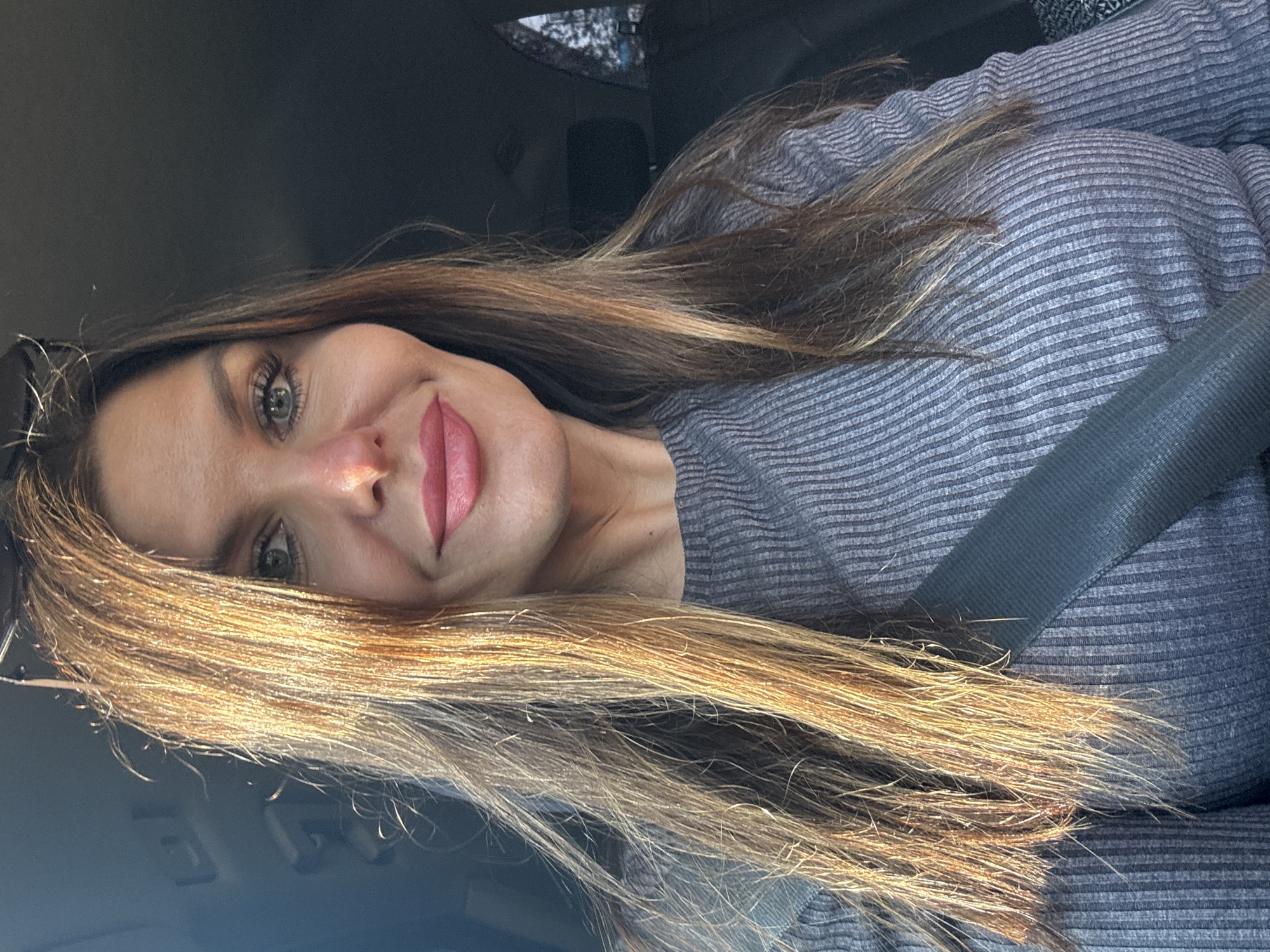 A woman with long, wavy, sun-kissed brown hair is captured in a close-up, waist-up portrait, likely taken inside a vehicle as indicated by the visible seatbelt and the interior of a car. She is wearing a ribbed, grey long-sleeved shirt. Her facial expression is one of a gentle smile, with her lips colored in a pink lipstick. Her blue eyes are looking directly at the camera. The lighting suggests it is daytime, with sunlight illuminating her hair from the left. The background is dark and slightly blurred, consistent with the interior of a car. No specific landmarks or context from Chaikovsky, Russia, are discernible in this image.
