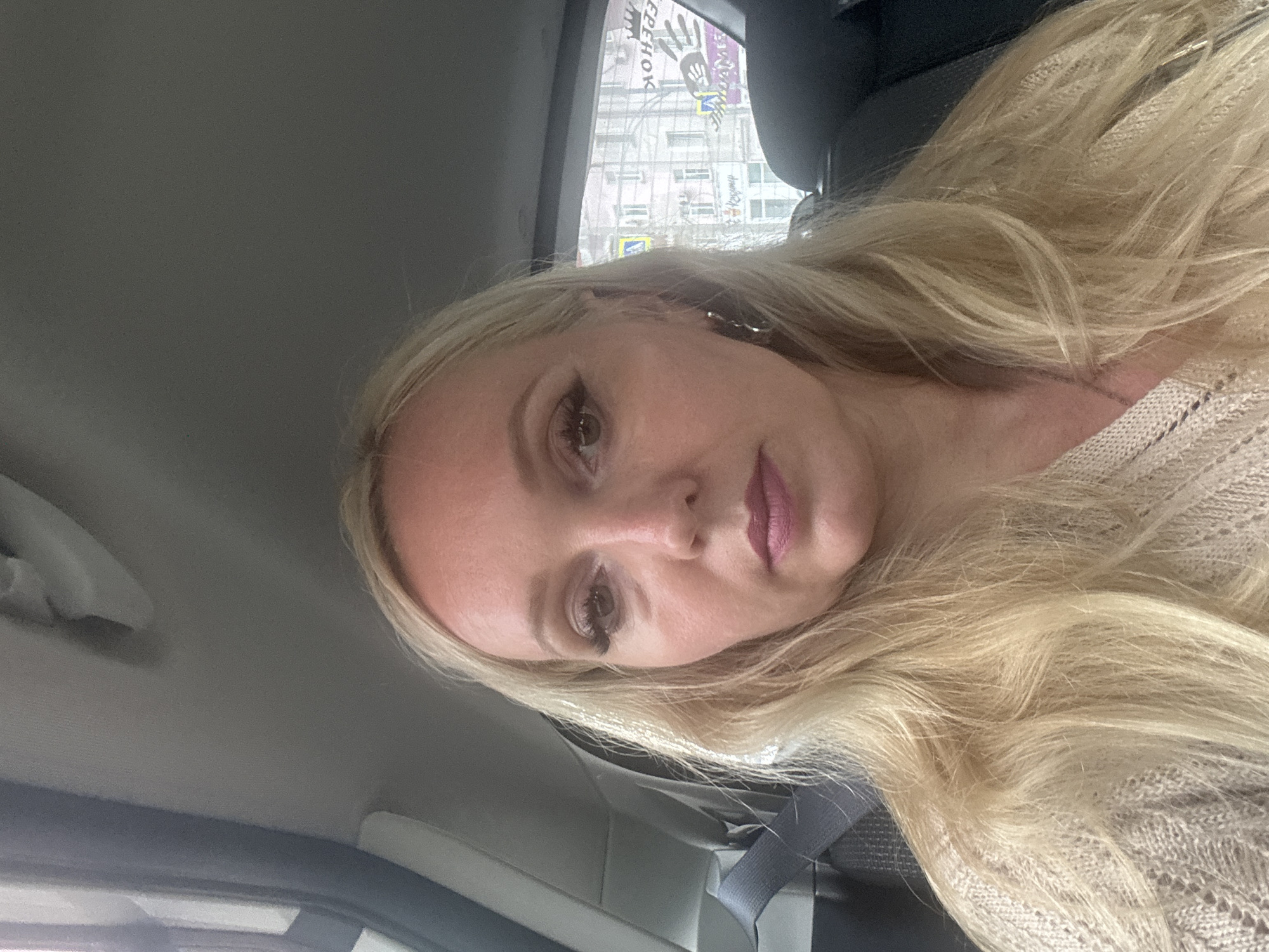 The image is a close-up, front-facing selfie of a blonde woman with fair skin, wearing makeup, including mascara, eyeliner, and lipstick. She is inside a vehicle, with a grey seatbelt visible on the left side of the frame. The interior of the car, specifically the ceiling and a portion of the backseat, is also visible. Through the car window, an urban environment is visible, featuring buildings with windows and what appear to be street signs or advertisements, some with Cyrillic lettering. The text on the visible signs suggests the location is Komsomolsk-on-Amur, Russia. The woman has long, blonde hair that is partially obscuring her shoulders and chest, and she is wearing a light-colored, textured sweater.