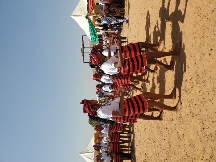 The scene depicts an outdoor event in Bar Gada, Nigeria, characterized by a clear blue sky and light brown dirt ground. Approximately fifteen to twenty individuals, predominantly women, are engaged in a synchronized performance or dance. They wear uniform attire: white short-sleeved tops, short skirts with horizontal red and black stripes, and matching red and black patterned headwraps. Most participants are barefoot. Several individuals in the foreground hold small, dark objects in their hands, consistent with props or instruments. In the background, two white canopy tents are visible; the tent on the right is draped with green and red fabric. A male individual, dressed in blue and white patterned clothing, stands near this tent. Other indistinct figures are present further in the background, observing the activity.