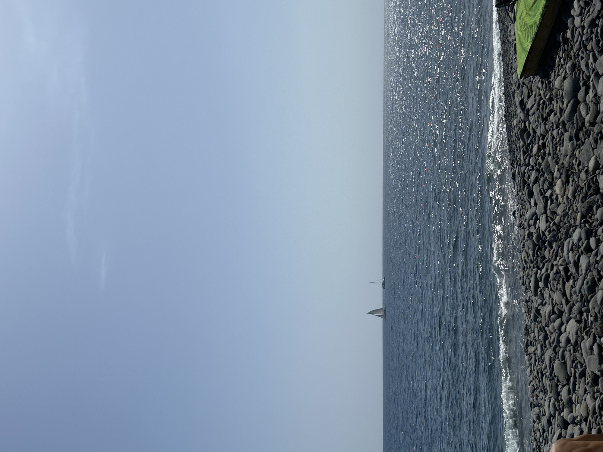 The image shows a view of the sea and a rocky beach in Câmara de Lobos, Portugal. The sky is clear blue. The sea is a darker blue with a few sailboats visible in the distance. The beach is made up of smooth, dark gray rocks. Waves are breaking on the shore. Part of a green object is in the top right. A small part of a person's skin is visible in the lower right corner.