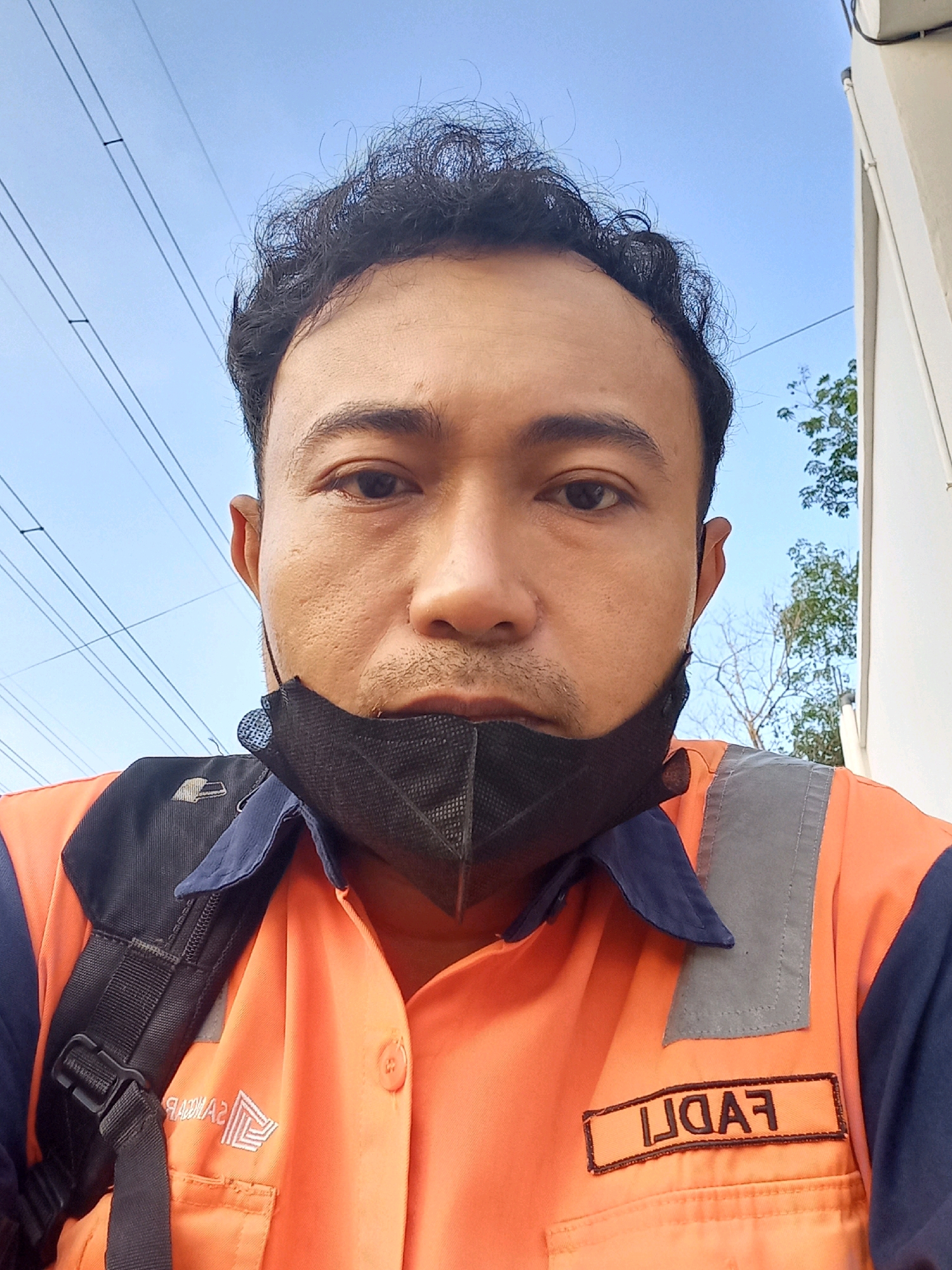 A close-up, chest-up, selfie-style, medium shot shows a man in Maburai, Indonesia. The man has dark, curly hair, brown eyes, and a light complexion. He is wearing a black face mask, an orange work shirt with a dark blue collar and sleeves, and a dark backpack. A reflective gray patch is visible on the right chest of his shirt, with a rectangular name tag embroidered with the name "FADLI" in black. The background consists of a clear blue sky, power lines, a white building, and some green foliage. The man appears to be outdoors during the daytime.