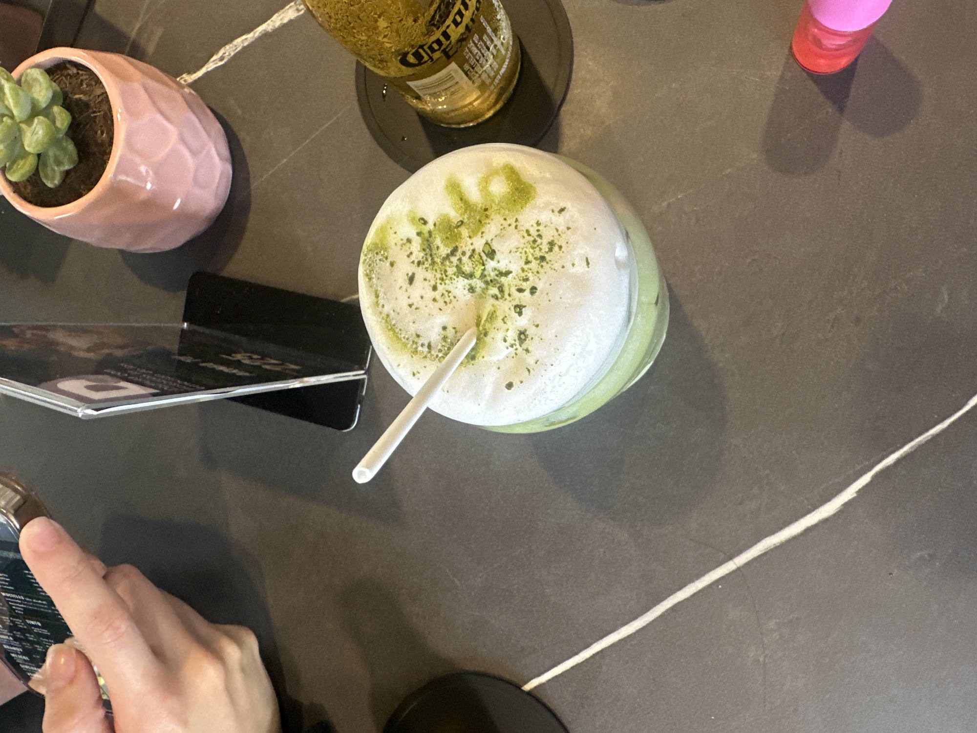 This image depicts a close-up, overhead view of a table in a modern establishment located in San Andrés Cholula, Mexico. The table surface is a dark grey material, possibly stone or laminate, traversed by a distinct white diagonal vein. In the center, a clear glass holds a light green matcha latte, topped with a thick layer of white foam and sprinkled with green matcha powder forming abstract patterns. A white drinking straw is inserted into the beverage. To the right of the latte, a bottle of Corona Extra beer rests on a circular black coaster. In the upper left quadrant, an individual's hands are visible holding a smartphone, whose screen displays indistinct digital content. Behind the smartphone, a small, black standing menu or sign holder is partially visible. Towards the upper right, a small succulent plant is potted in a pink, geometrically faceted ceramic container. A small cylindrical object, red at the top transitioning to pink, is partially visible in the bottom right corner of the frame. The lighting is ambient, indicating an indoor setting consistent with a cafe or bar.