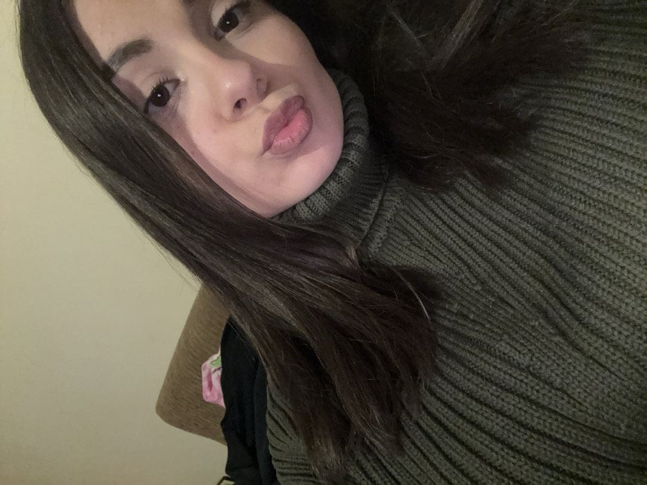 A close-up, selfie-style image captures a young female indoors in Radomir, Bulgaria. The subject has long, dark brown hair and dark eyes, and is wearing an olive-green ribbed turtleneck sweater. Her head is tilted slightly to her left, and her lips are puckered in a "kiss" expression, directed towards the camera. The background reveals a light-colored wall and the upper portion of a tan, upholstered furniture item, possibly a couch or headboard, upon which dark fabric and a pink patterned item are visible.