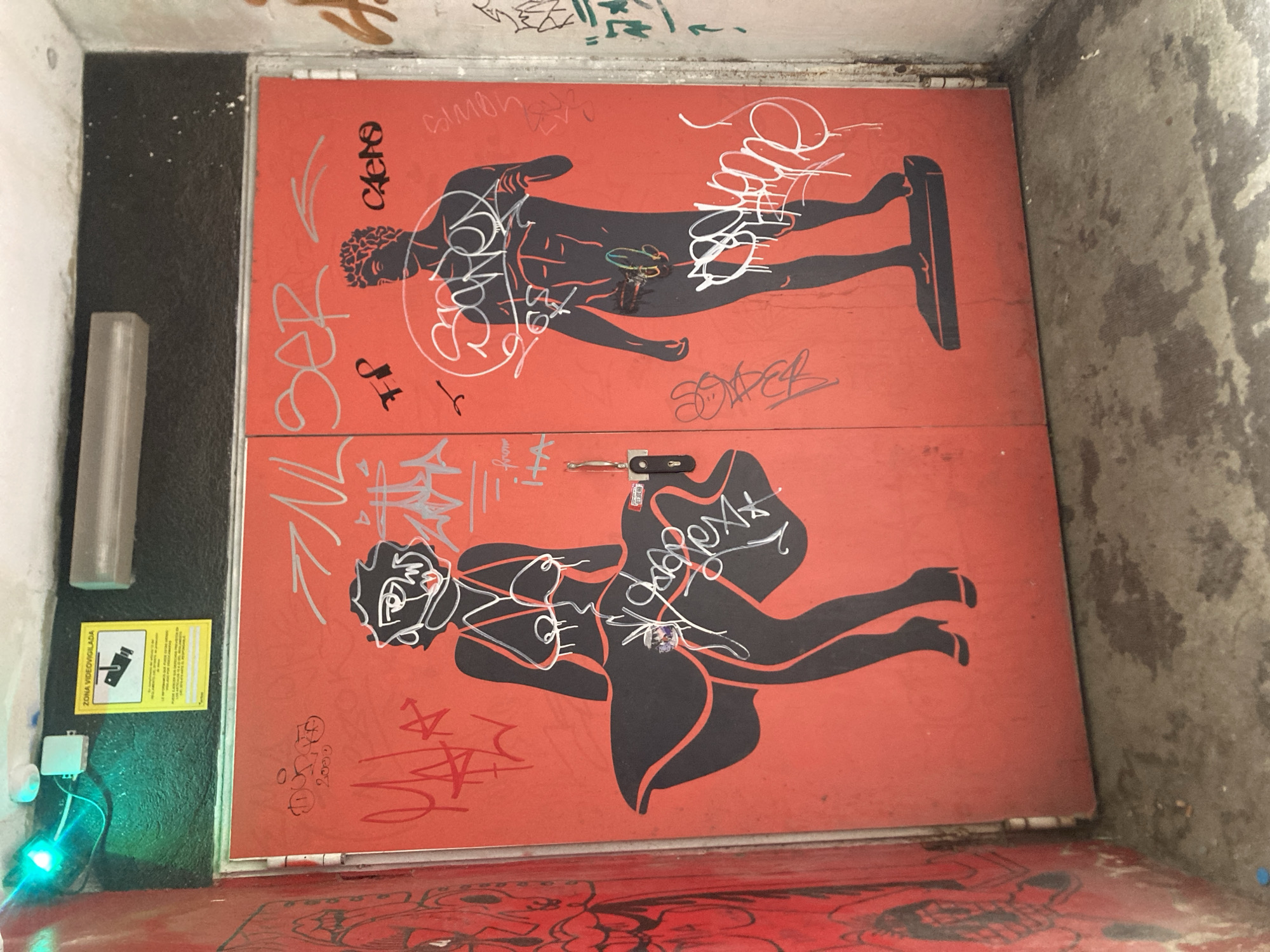 The image shows two red doors covered in graffiti, located in Barcelona, Spain. On the left door, a stylized black silhouette of Marilyn Monroe is depicted, her dress billowing around her legs. She is wearing high heels. On the right door, a black silhouette of a muscular man is shown, with graffiti scribbled over his body and around him. His genitalia are also depicted and graffitied. Above the doors, a yellow sign with a camera symbol and Spanish text "ZONA VIDEOGUILADA" (Surveillance Zone) is visible, along with a green light. The environment appears to be an alleyway or entryway with concrete flooring and grimy walls.