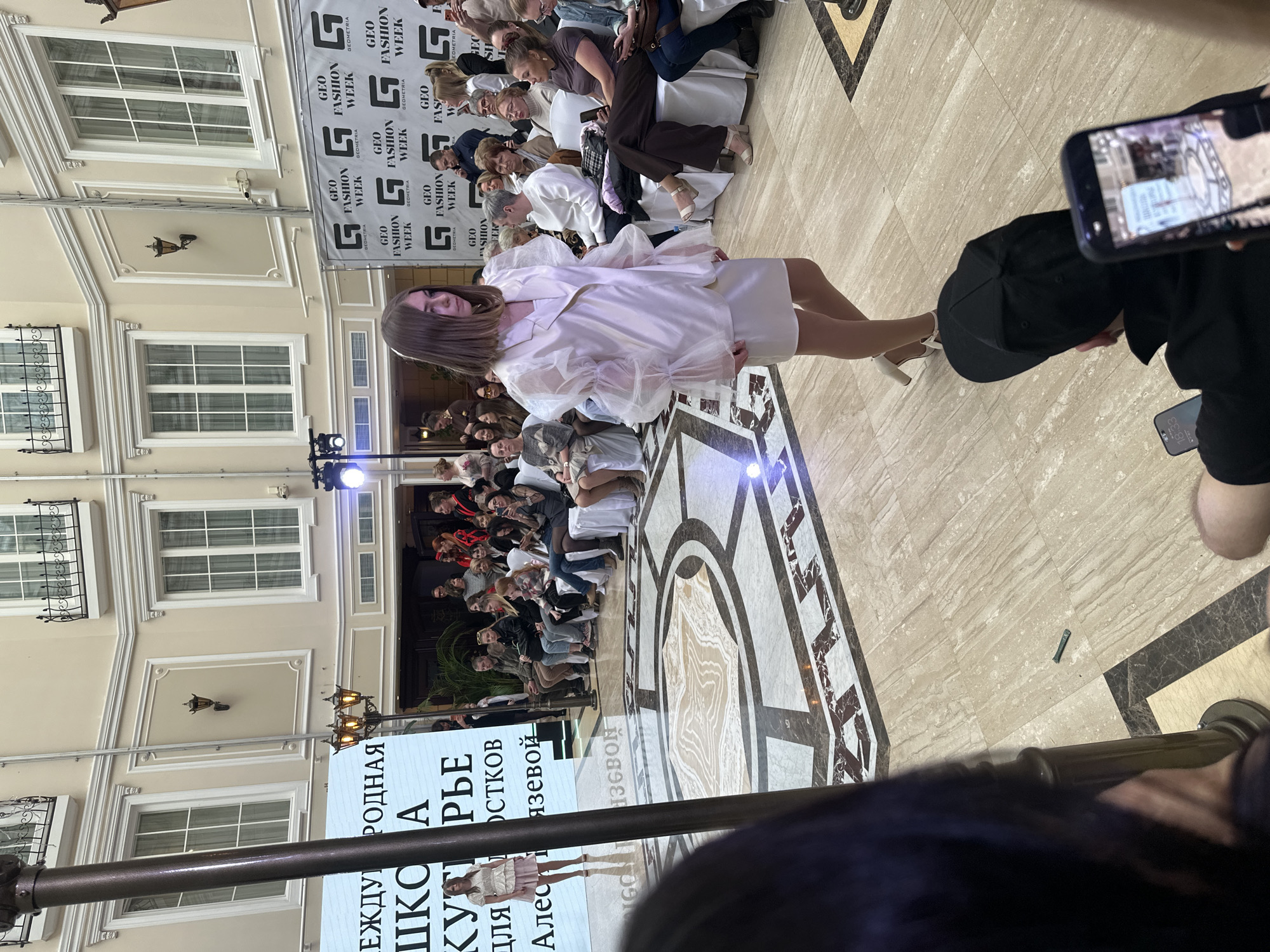 Here is a forensic description of the image:
**Scene:** The image captures a fashion show. The model walks the runway while spectators watch. Banners advertising "GEO FASHION WEEK" and a promotional board for "РОДНАЯ ШКОЛА КУТІ РЬЕ" are displayed in the background. The building exterior visible through the back shows an ornate style.
**People:** A model is on the runway, wearing a white outfit. There are several spectators on both sides of the runway. In the foreground, the phone of a spectator is capturing video of the event.
**Objects:** A runway, banners, chairs, a phone.
**Actions:** The model is walking the runway. Spectators are watching and taking photos/videos.
**Environment:** The event seems to be taking place inside a building with marble tile flooring and ornate architectural details.