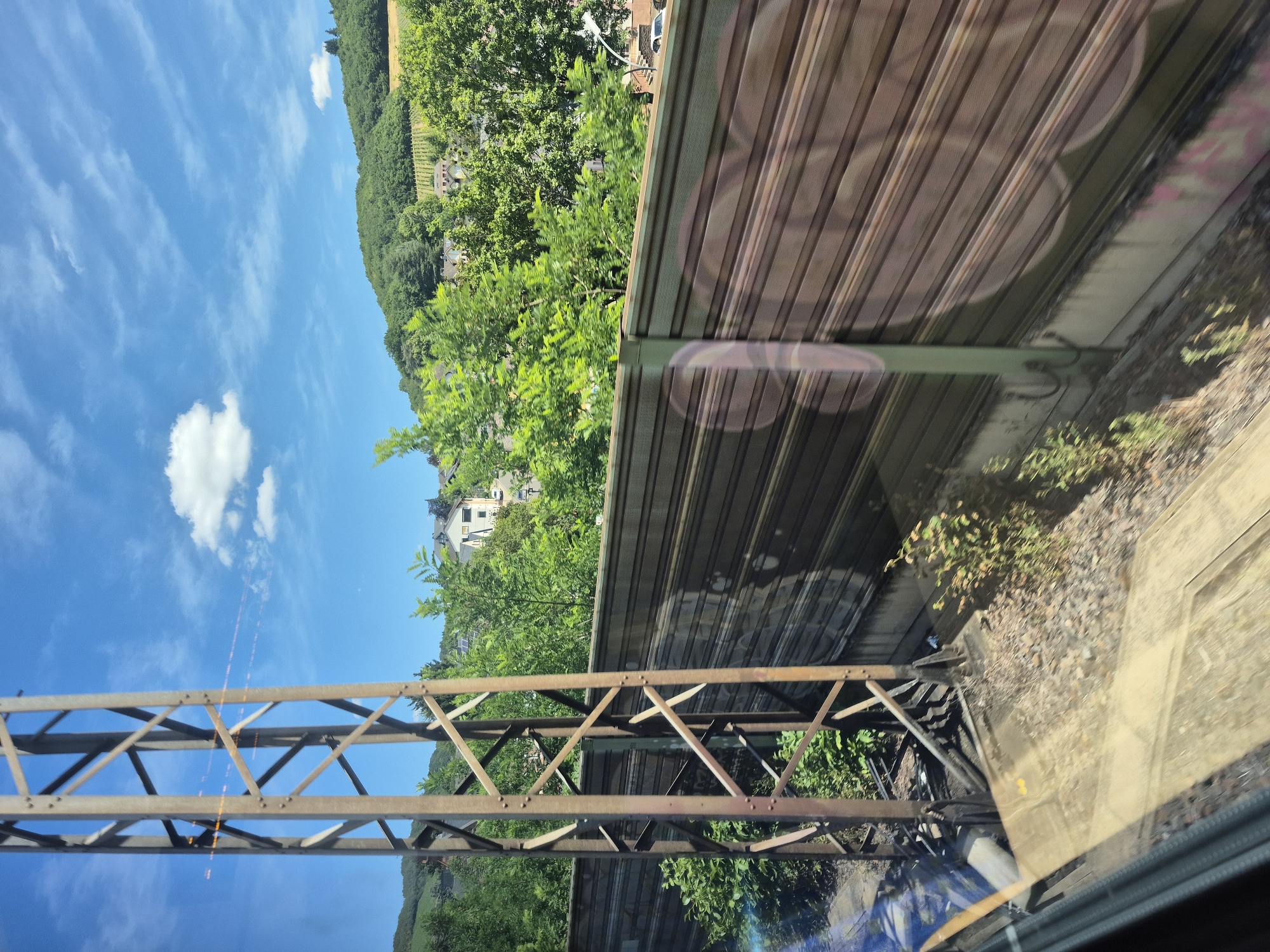 The image depicts a view from a train traveling through the city of Kobern-Gondorf, Germany. A tall, rusted metal pylon is visible on the left side of the frame, positioned close to the train. A long, horizontal fence, likely a sound barrier, runs along the right side of the track. Beyond the fence, a hillside covered in lush green trees and vineyards is visible. Several buildings, typical of a German town, are scattered among the trees on the hillside. The sky is a bright blue with scattered white clouds, suggesting fair weather. The overall scene conveys a sense of travel through a scenic, rural area of Germany.