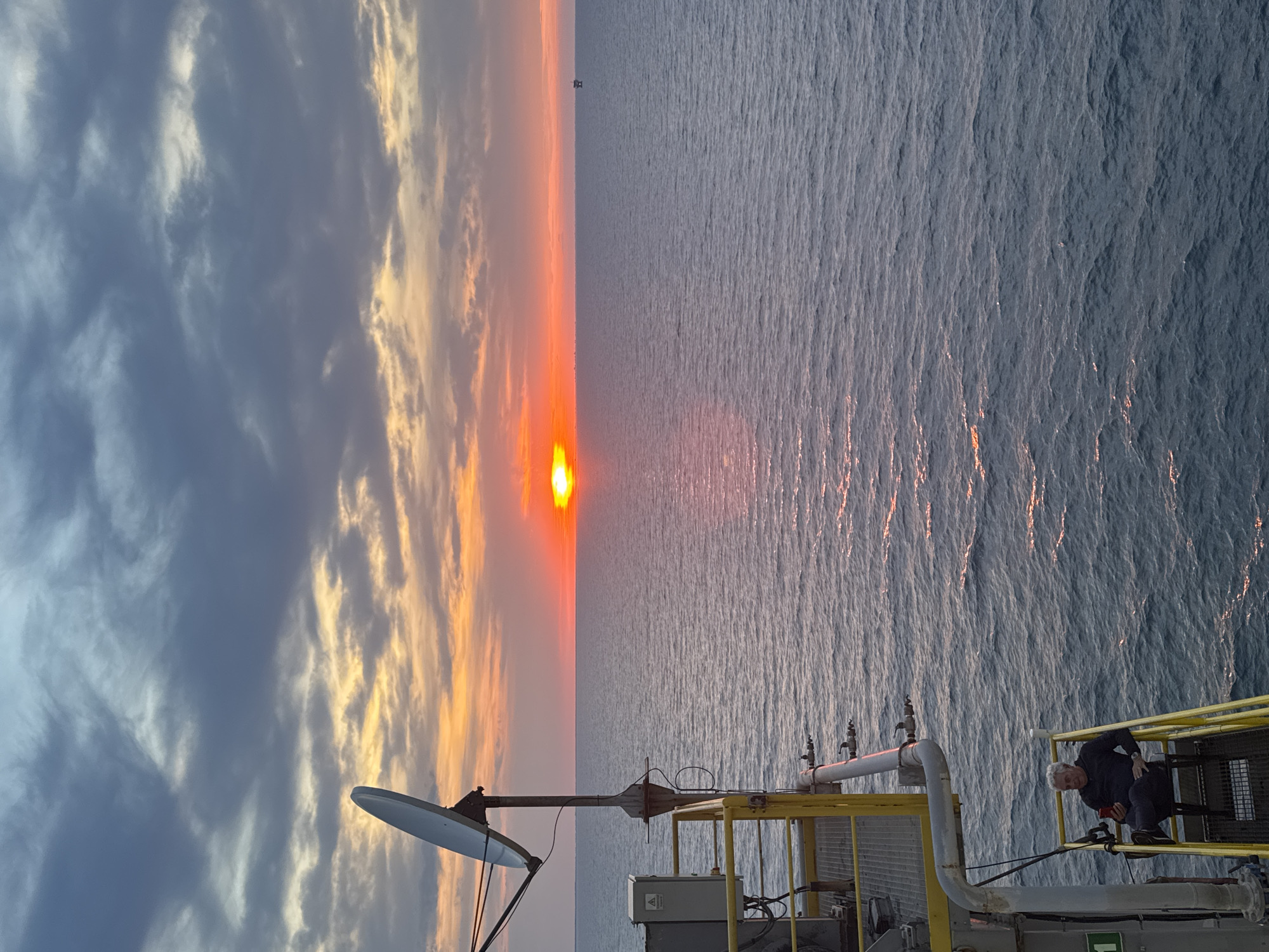 A wide shot captures an offshore platform's edge during a sunset over the ocean. The sky displays a gradient of colors, from dark gray clouds at the top to bright orange and yellow hues near the horizon where the sun is setting. A large satellite dish is mounted on the platform structure, which is painted yellow and gray. A man, seated on a chair with a red object in his hand, is visible on a yellow metal staircase attached to the platform. The ocean surface is textured with small waves reflecting the sunset's light. In the distance, a small structure, possibly another platform or buoy, is visible on the horizon. The location is Unknown, Italy.