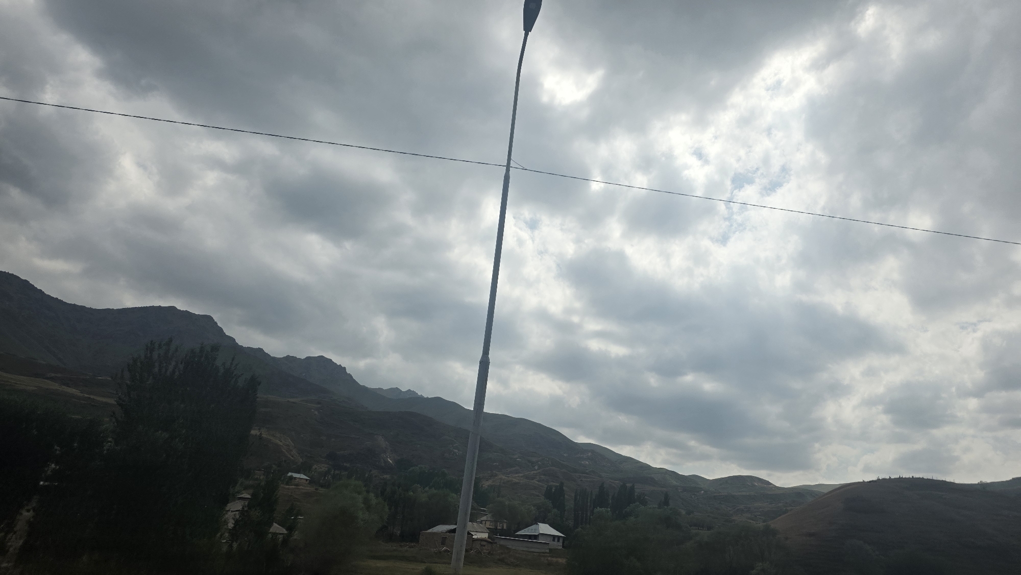 The image shows a cloudy sky over a mountainous landscape in the vicinity of Taldyk, Kyrgyzstan. A tall, slender lamppost stands prominently in the center of the frame, leaning slightly. A thin wire stretches horizontally across the upper portion of the image, passing behind the lamppost. In the foreground and mid-ground, rolling hills are covered with vegetation, and several small, scattered buildings are visible. The mountains in the background are dark and rugged, with visible ridges and slopes. The overall lighting suggests an overcast day. No people or animals are discernible in the scene.