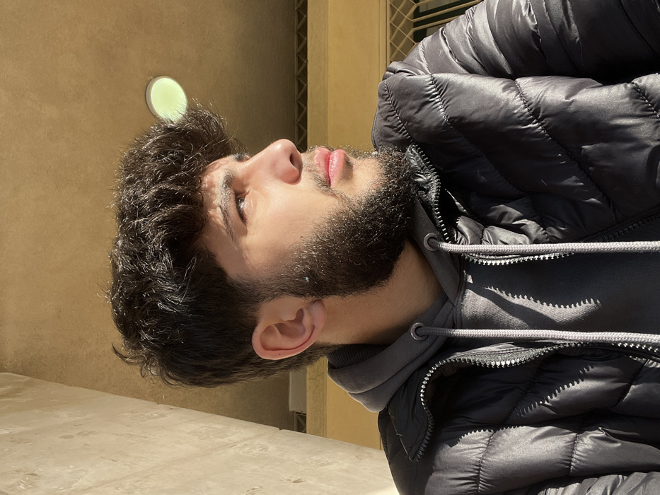 A young man with dark, curly hair and a trimmed beard looks upwards. He wears a grey hooded sweatshirt beneath a black puffer jacket. The background features a beige wall with a circular light fixture and a metal grate. Sunlight highlights his face and casts shadows on the jacket. The image is rotated counterclockwise.