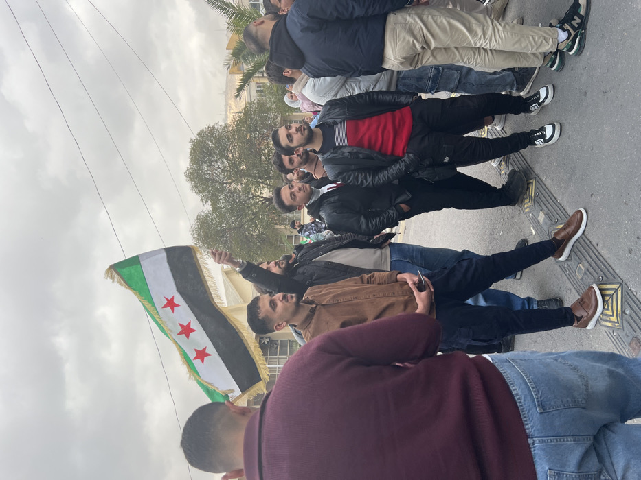 The image displays an outdoor gathering of approximately 15 to 20 individuals, primarily young men, situated on a paved street in Al-Qanawat Municipality, Syria. The scene is illuminated by an overcast sky. In the mid-ground, a man wearing a dark jacket holds a flag horizontally; the flag consists of three horizontal stripes (green, white, black from top to bottom) and three red five-pointed stars centered on the white stripe, bordered by a gold fringe. In the left foreground, a man is seen from the back, wearing a maroon sweater and blue jeans. To his right, a man in a brown collared shirt and dark cargo pants stands on a yellow tactile paving block while holding a smartphone. Other individuals in the mid-ground and background are dressed in various casual attire, including dark jackets, sweaters, and jeans. The ground is paved, with additional yellow tactile paving blocks visible. Overhead, multiple power lines crisscross the sky. The background features light-colored urban buildings, deciduous trees, and a palm tree, with a distant elevated area or hill partially visible. The individuals are standing and appear to be observing the scene, with no discernible active interactions or specific actions beyond their assembly.