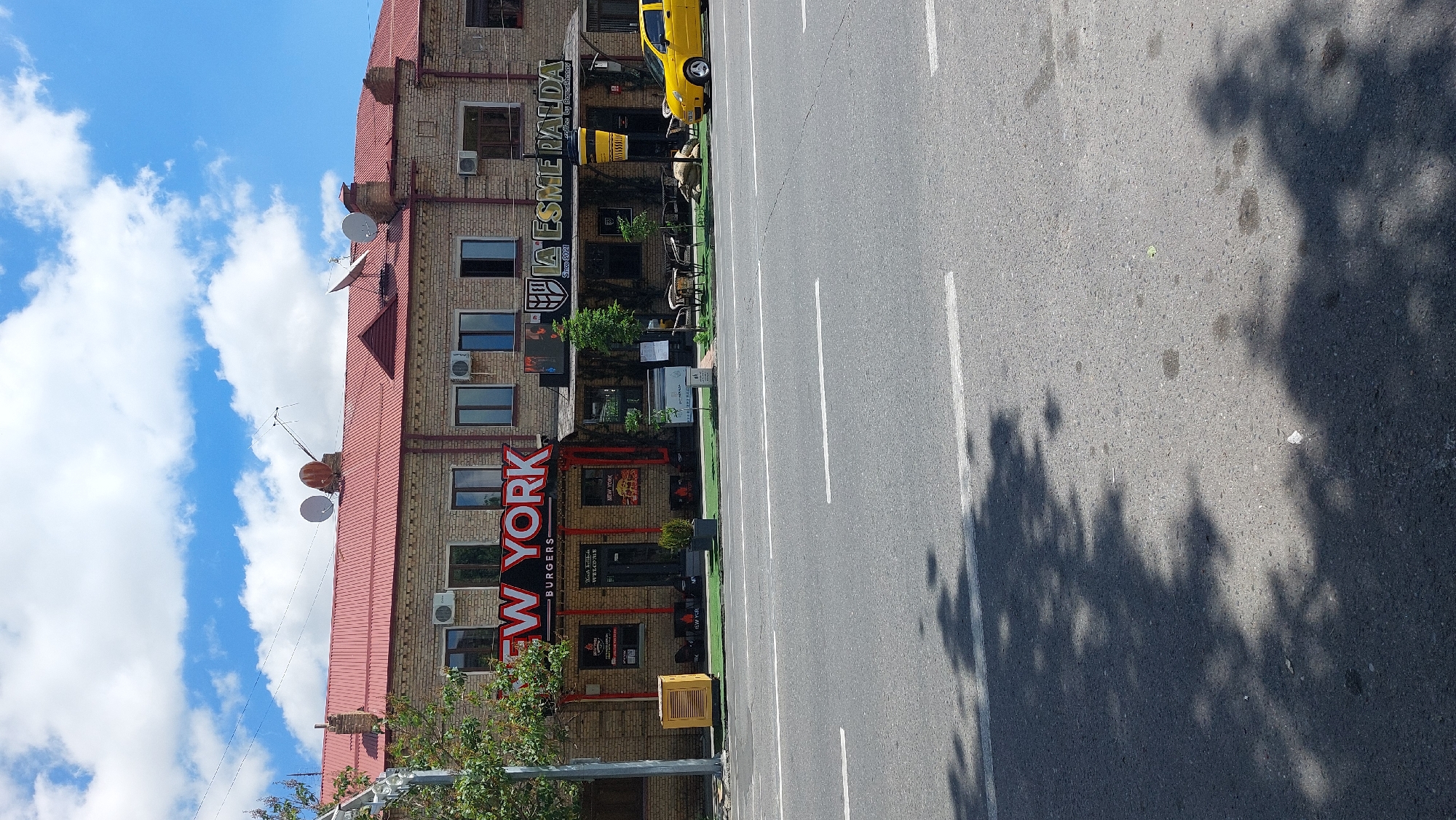 The image shows a street in Samarkand City, Uzbekistan, featuring two businesses on the left: "New York Burgers" and "La Esmeralda." A yellow vehicle is parked in front of the latter. The street has white lane markings, and shadows from trees are cast on the asphalt. The building facades are brick, with visible windows and air conditioning units. The sky is blue with scattered clouds.