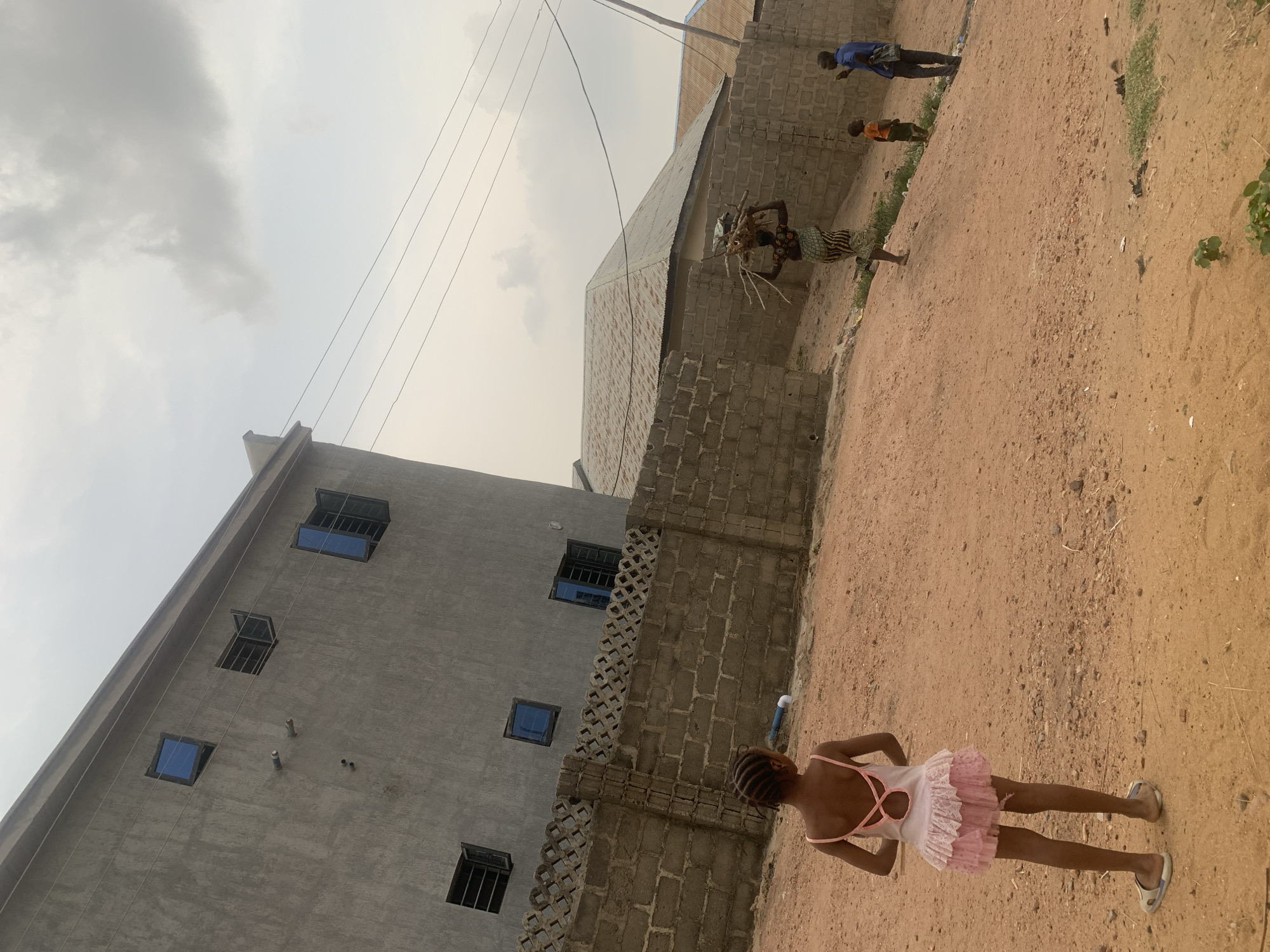This outdoor image, likely captured in Lokoja, Nigeria, depicts an everyday scene on an unpaved, reddish-brown dirt path under an overcast, bright sky.
In the foreground, a young girl stands facing away from the camera, looking towards the left side of the frame. She has dark braided hair and wears a light pink, sleeveless dress with ruffles and a criss-cross design on the back. She is wearing sandals.
In the middle ground, slightly to the left, a person, likely a woman, is seen from behind, carrying a substantial bundle of dry firewood or branches balanced on their head. They are dressed in a patterned garment, possibly a wrapper skirt. Further back on the dirt path, two other individuals are visible: one person in a blue top and dark trousers, and a smaller child wearing an orange top.
The setting features several buildings. On the left, a multi-story concrete building, appearing unpainted or partially constructed, has multiple small, square windows fitted with blue glass and security bars. Power lines are visible stretching across the upper portion of the sky, passing near this building. Another building with unplastered concrete block walls and a corrugated metal roof is visible behind the person carrying firewood. The ground is predominantly bare earth with scattered pebbles and sparse green vegetation.
The image portrays a routine daytime scene, without any visible text or discernible strong emotions from the subjects.