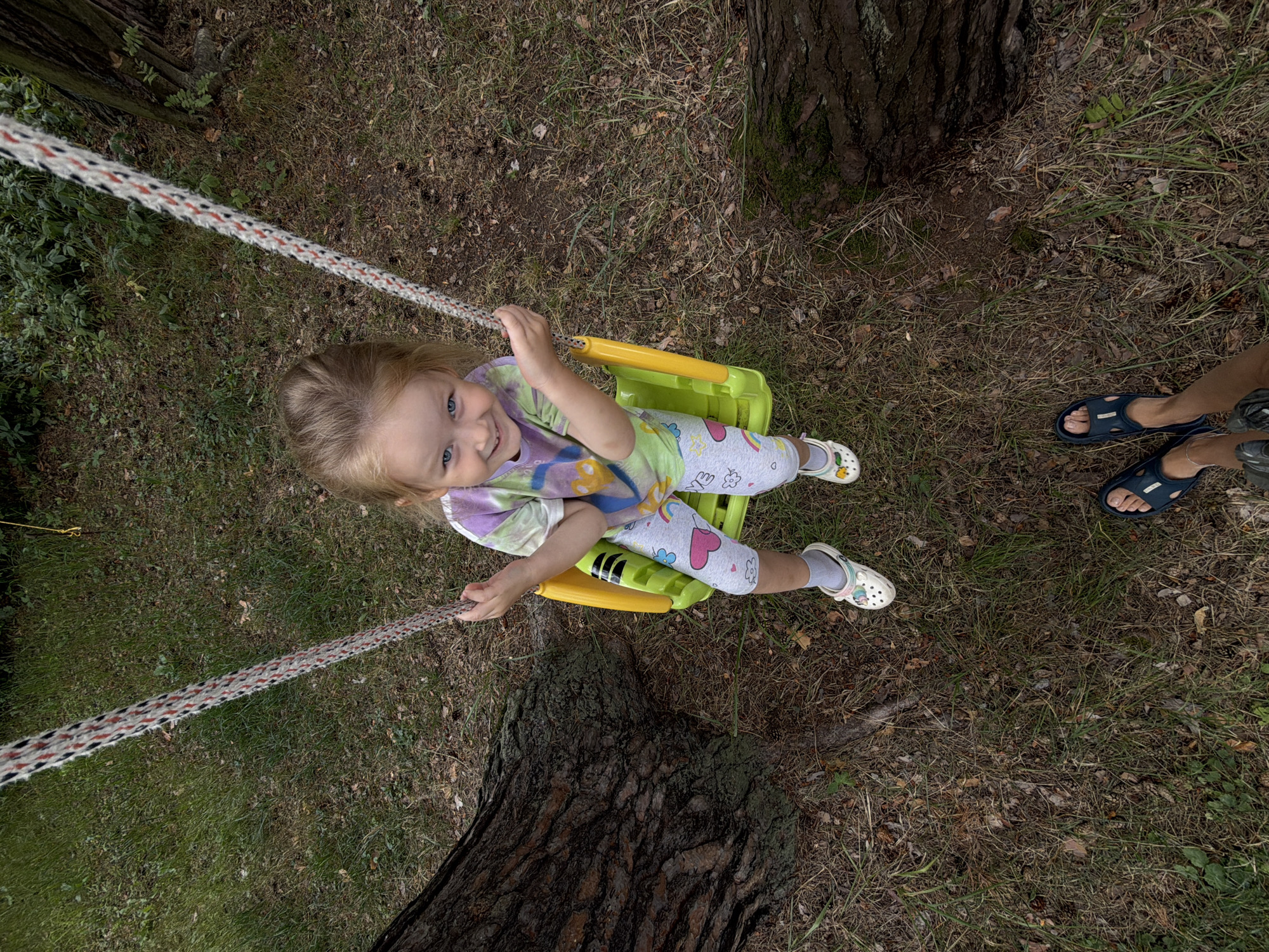 A young girl with blonde hair is sitting on a yellow swing in a wooded area. She is wearing a colorful t-shirt and leggings with heart and rainbow designs. She is smiling and looking at the camera. The swing is attached to ropes that are hanging from a tree. The ground is covered in grass, leaves, and pinecones. The perspective of the shot is from someone standing in front of the swing, looking up at the girl. Their feet, wearing dark sandals, are visible at the bottom of the frame.