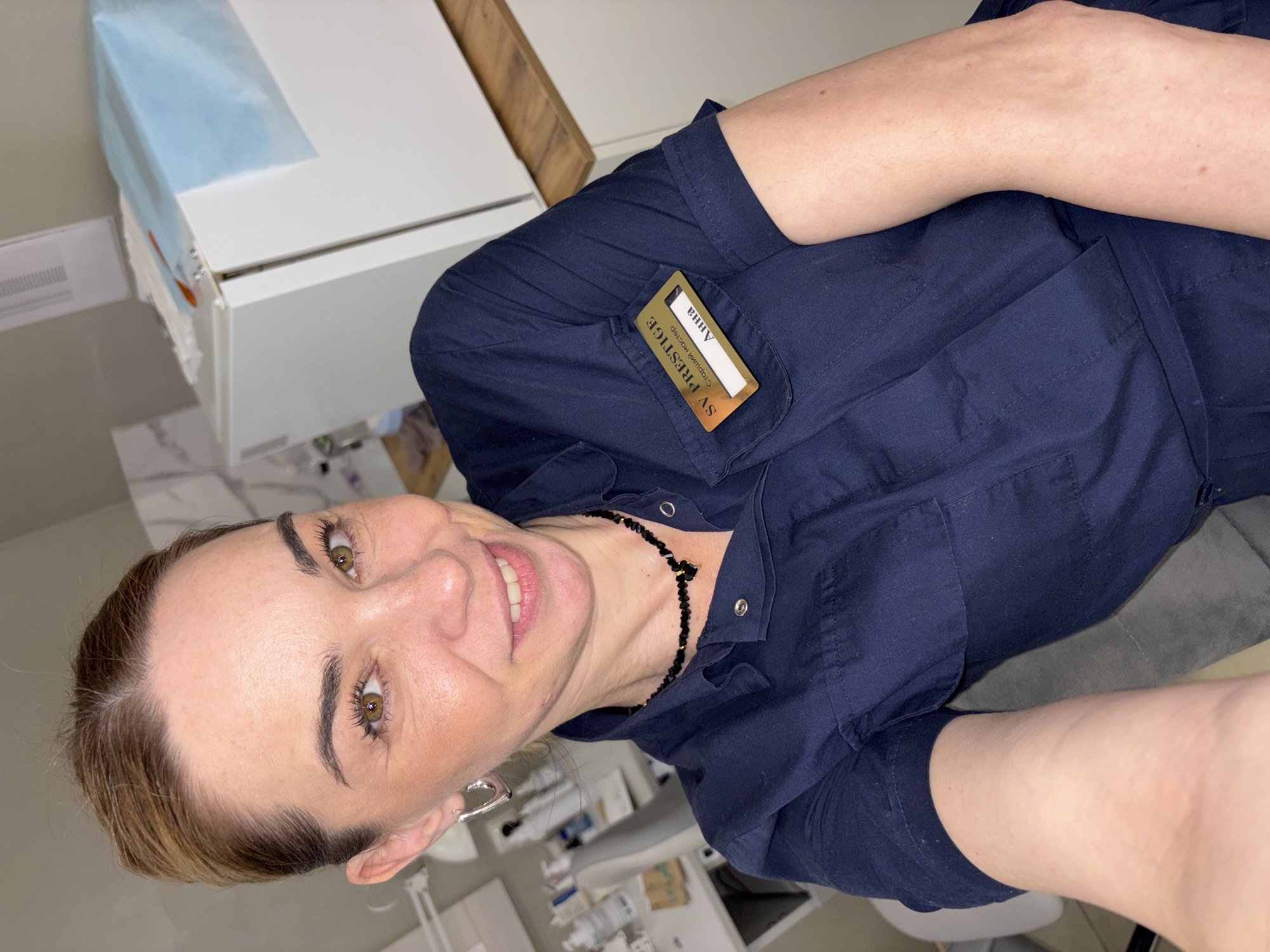 A woman with brown hair and eyes is facing the camera with a smile. She is wearing a dark blue button-up shirt with a name tag that reads "SV Prestige Анна" in Russian. She is also wearing a black beaded necklace. The background appears to be a medical or beauty-related environment with white cabinets and various bottles and equipment. Based on the information on the name tag, the location is likely in Vsevolozhsk, Russia.